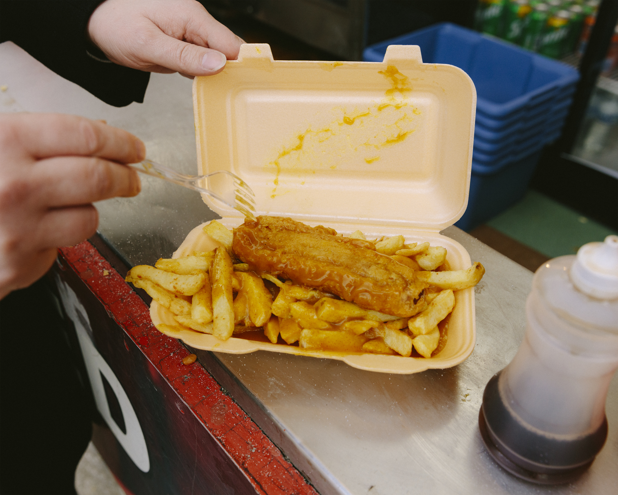 A battered sausage and chips dripping with curry sauce in a takeaway box.