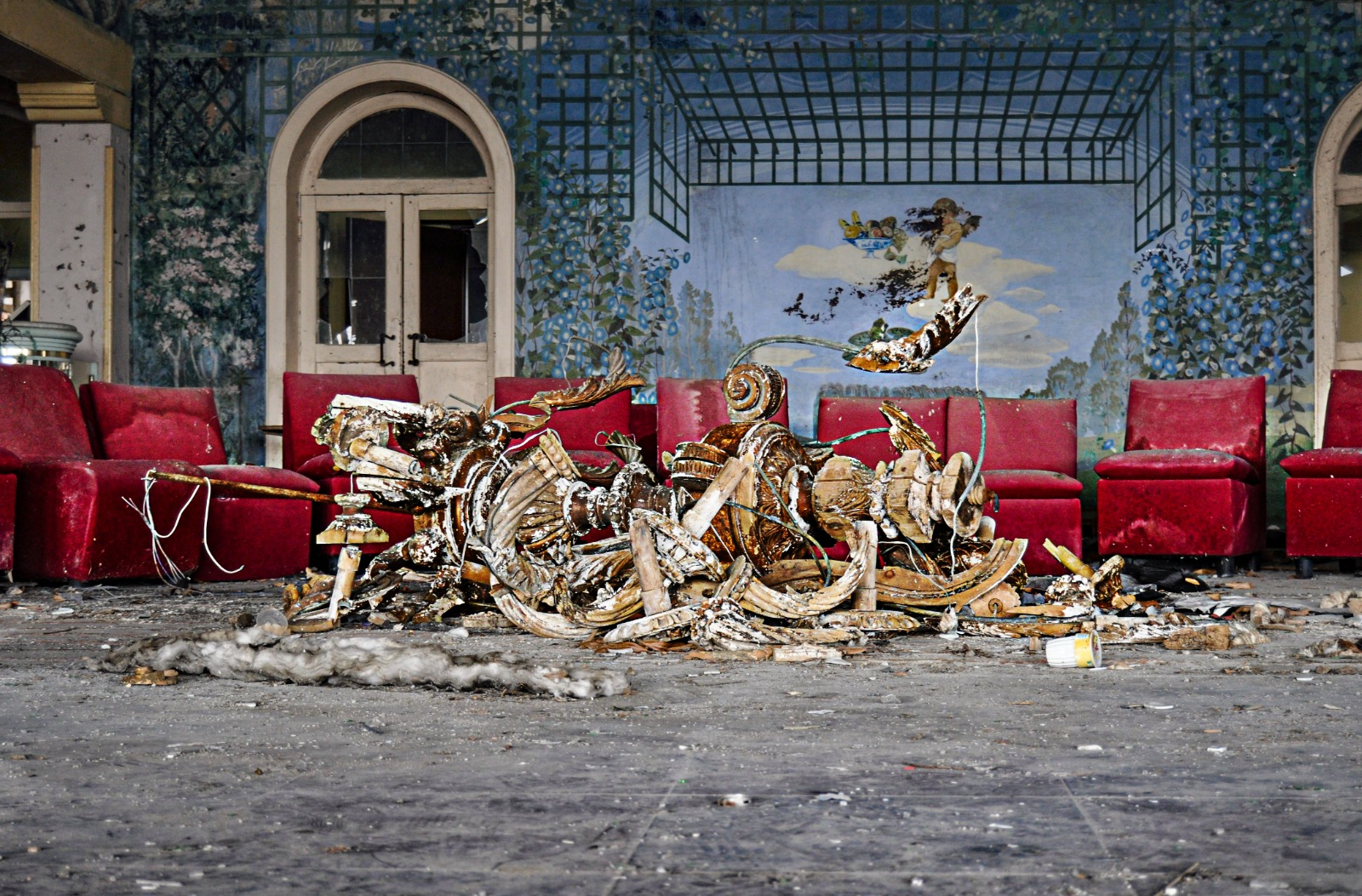 Italian Club Urbex, Woodpecker – The walls of the club are painted with images of natural landscapes. The space is filled by red chairs and there's a large and elaborate golden object lying in pieces in the middle.
