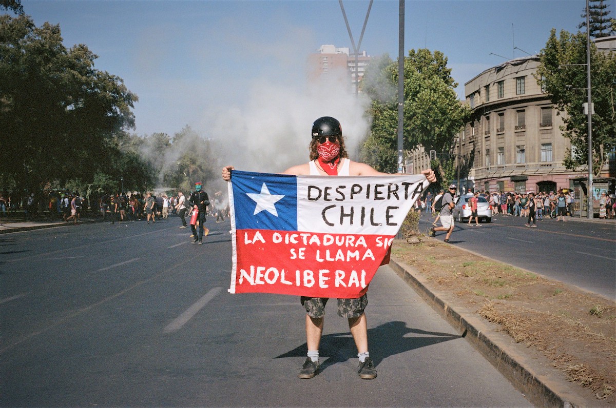 Chile protests – protester with a helmet, sunnies and a red bandana on their face, holding a sign while people are gathered behind them.
