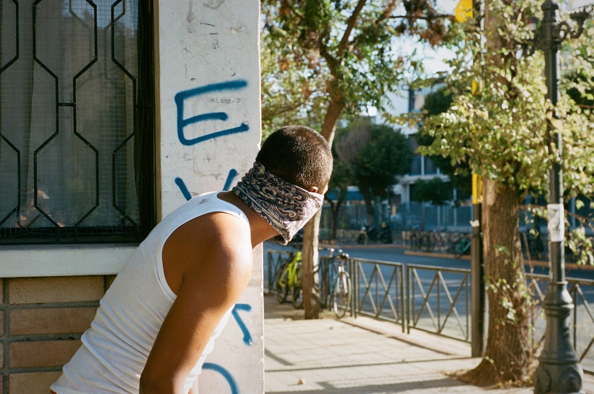 Chile protests – man in a white tank top and a bandana which covers his face, peaking over a building onto an empry street.