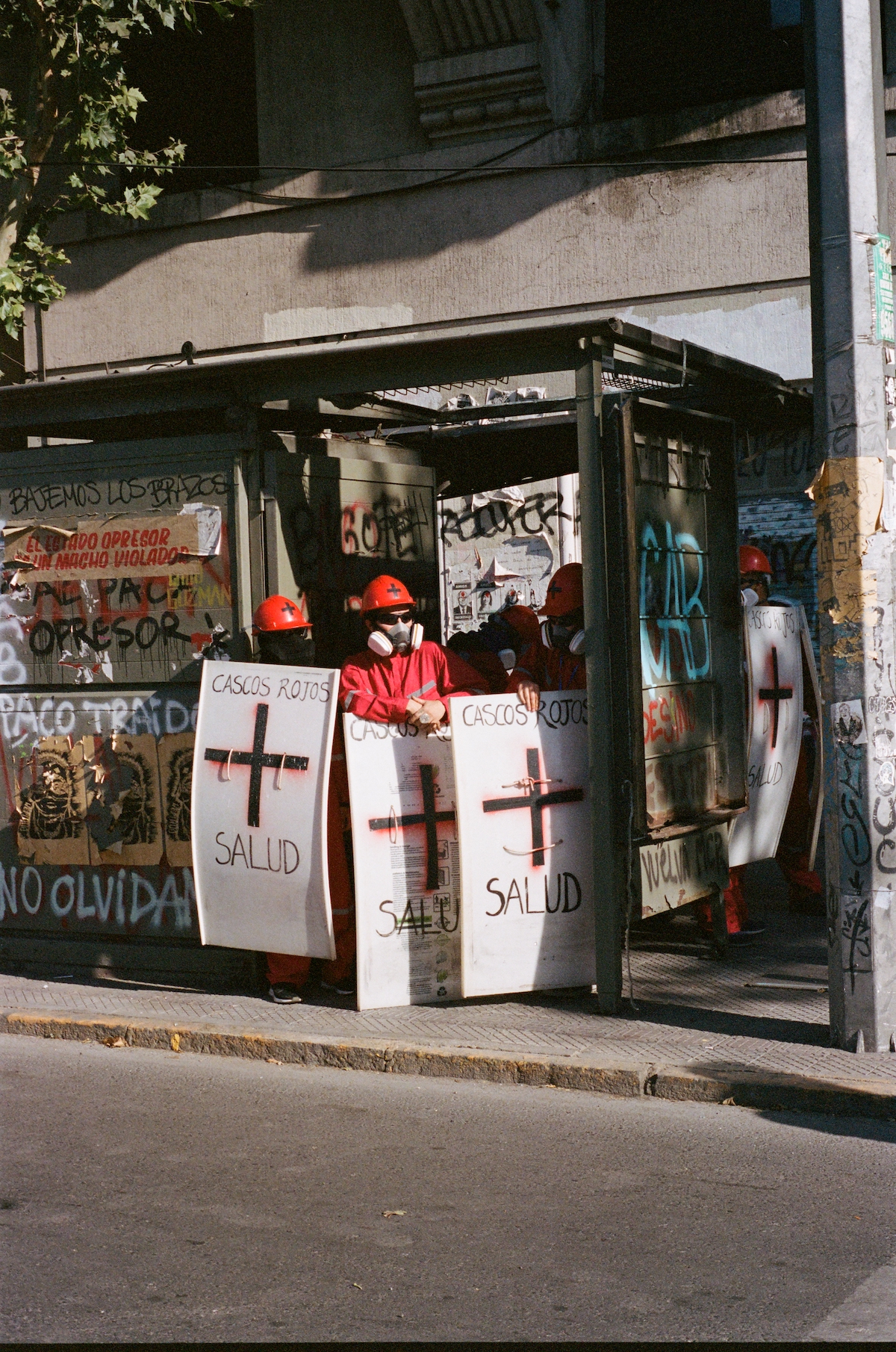 Chile protests – people wearing red suits, red helmets, sunglasses, gas masks and carrying white anti-riot shields with crosses, reading red cross.