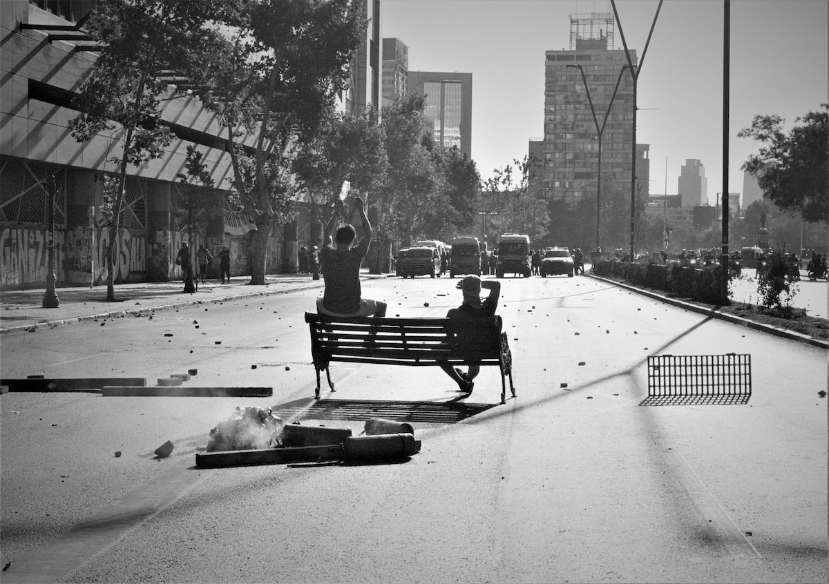 Chile protests – black and white picture of two guys sitting on a bench in front of a police barricade.