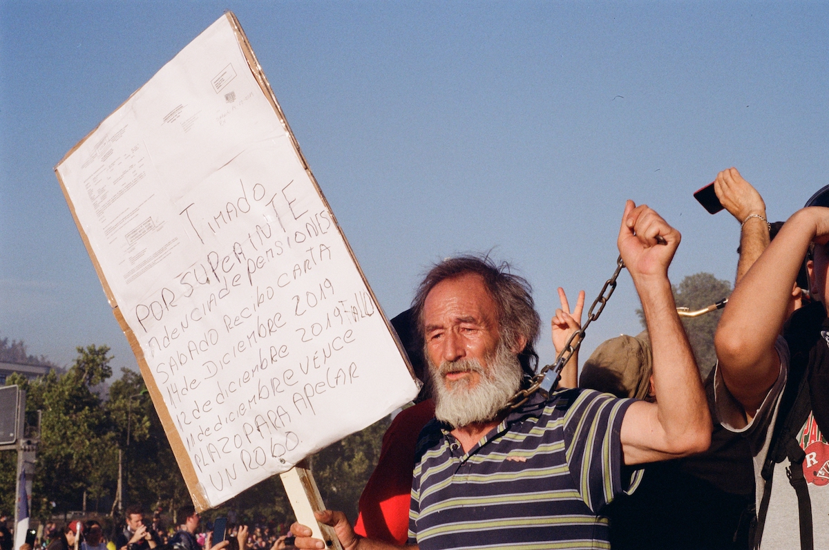 Chile protests – Old man with long hair and a beard, holding a sign in one hand and a chain wrapped against his neck in the other.