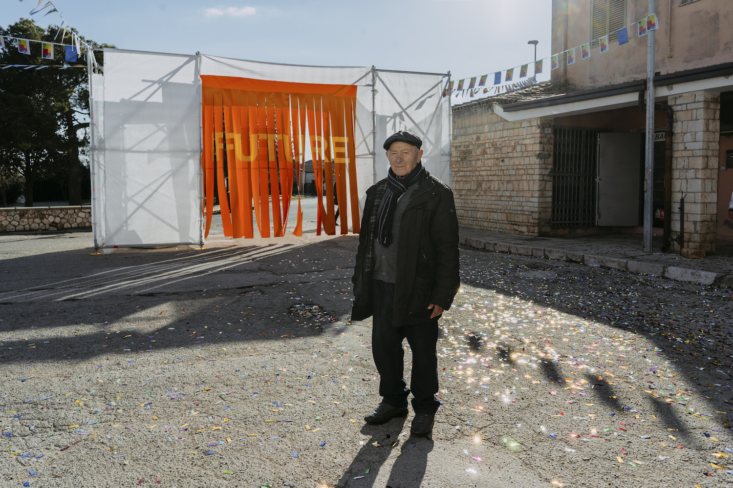 Michele Battilomo, De-population, Basilicata – Old man wearing a flat cap and a winter jacket standing in a courtyard covered in confetti in front of an archway made of scaffolding with