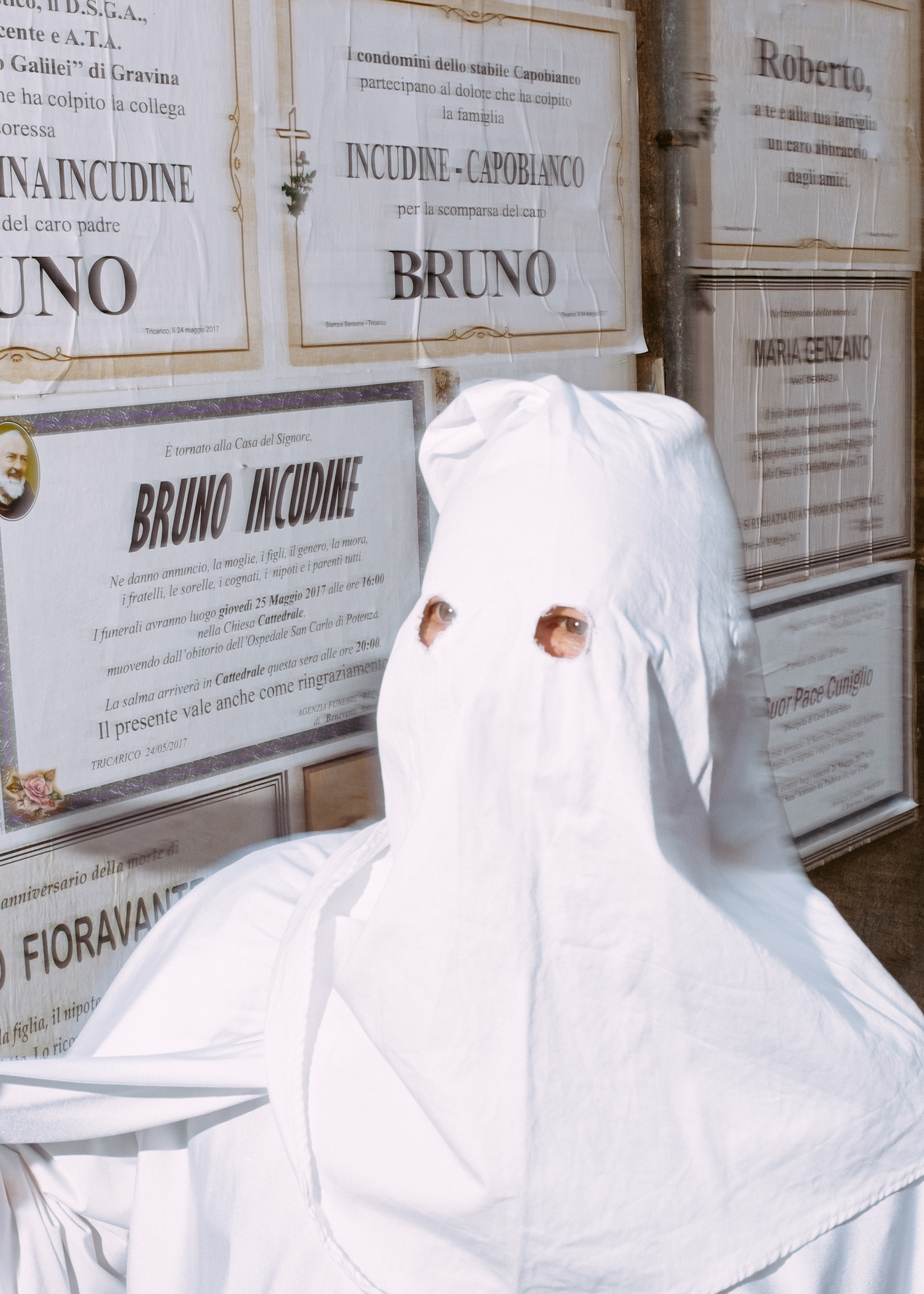 Michele Battilomo, De-population, Basilicata – Man wearing white cotton sheets covering his face with two holes for his eye, standing in front of a bulletin board with ads from people who've recently passed.