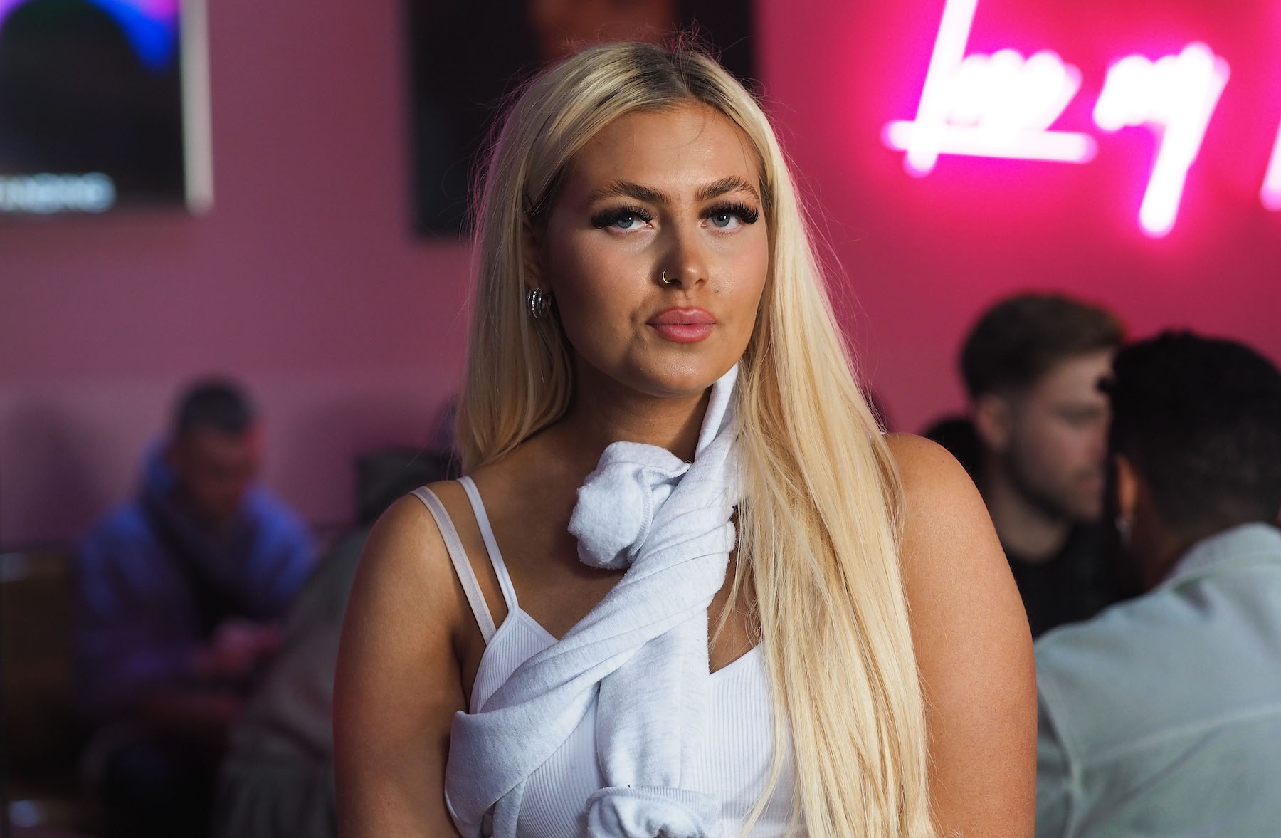 Eine junge Frau mit blondierten langen Haaren sitzt vor einem violetten Hintergrund in einer Bar