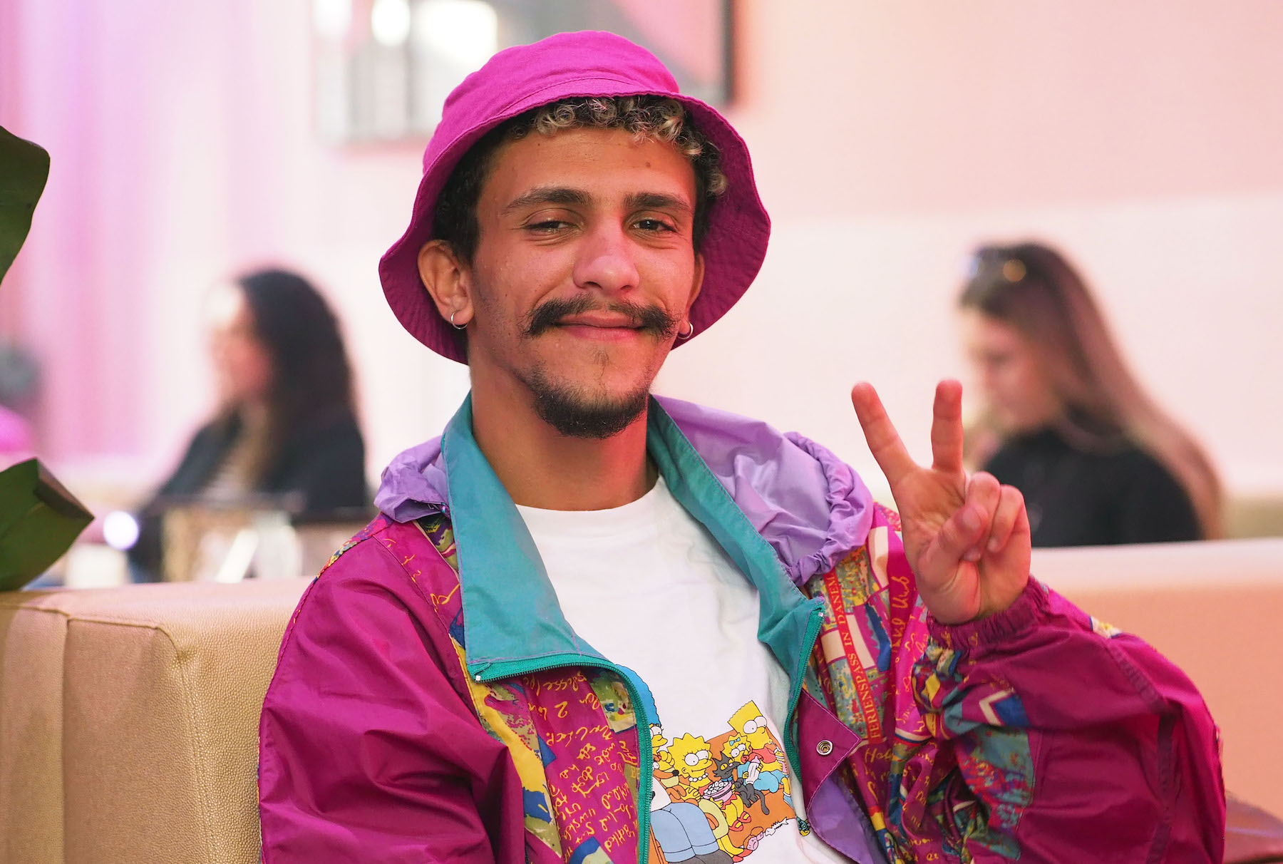 Ein junger Mann mit violettem Fischerhut und einem Simpsons-T-Shirt macht müde blickend ein Peace-Zeichen in die Kamera