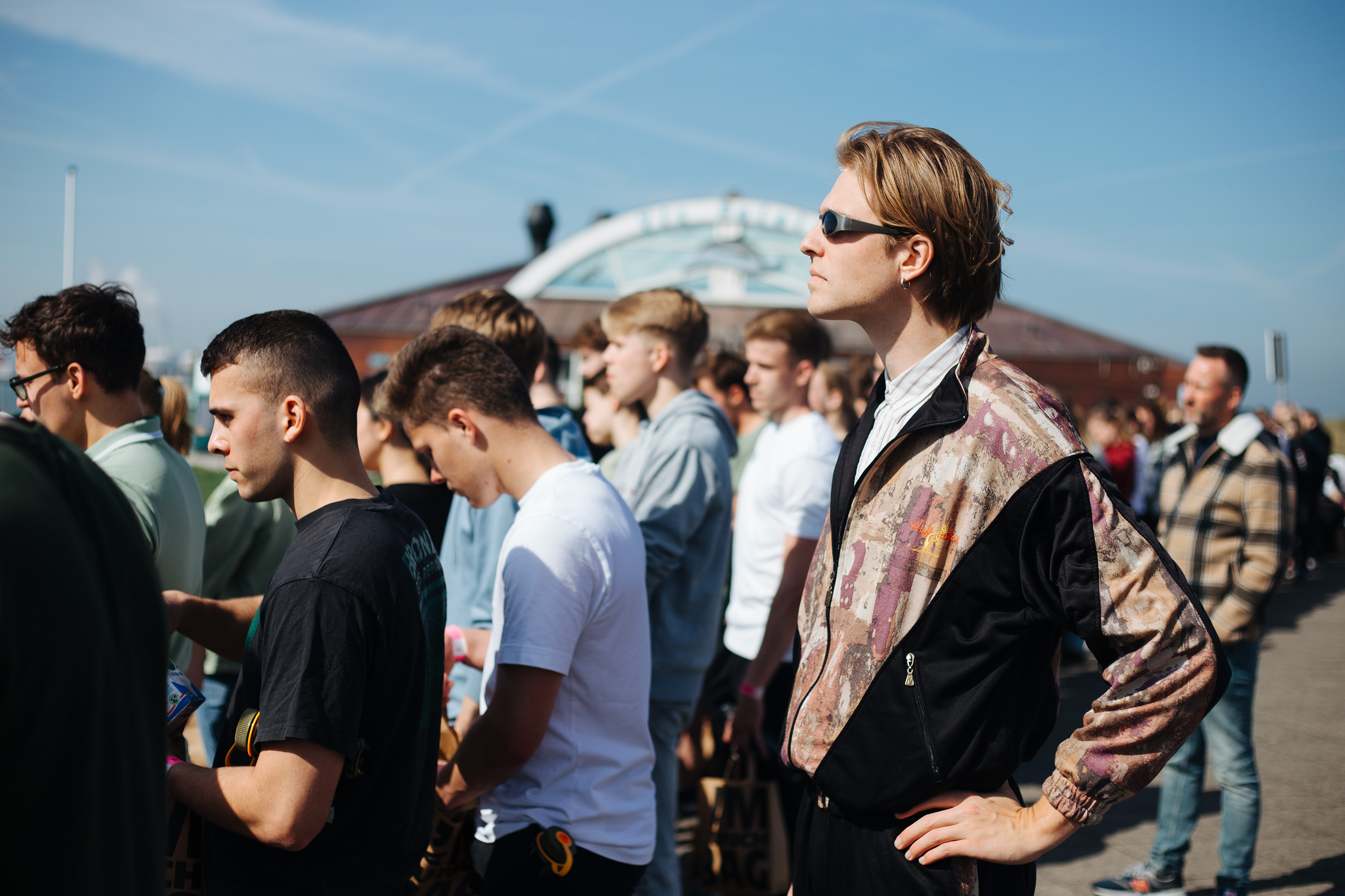 Army recruitment, Fort 1881 – The author, a blond man with long hair, wearing a black, mauve and beige 80s windbreaker and sunglasses, listening to a speech in a crowd of mostly young men.