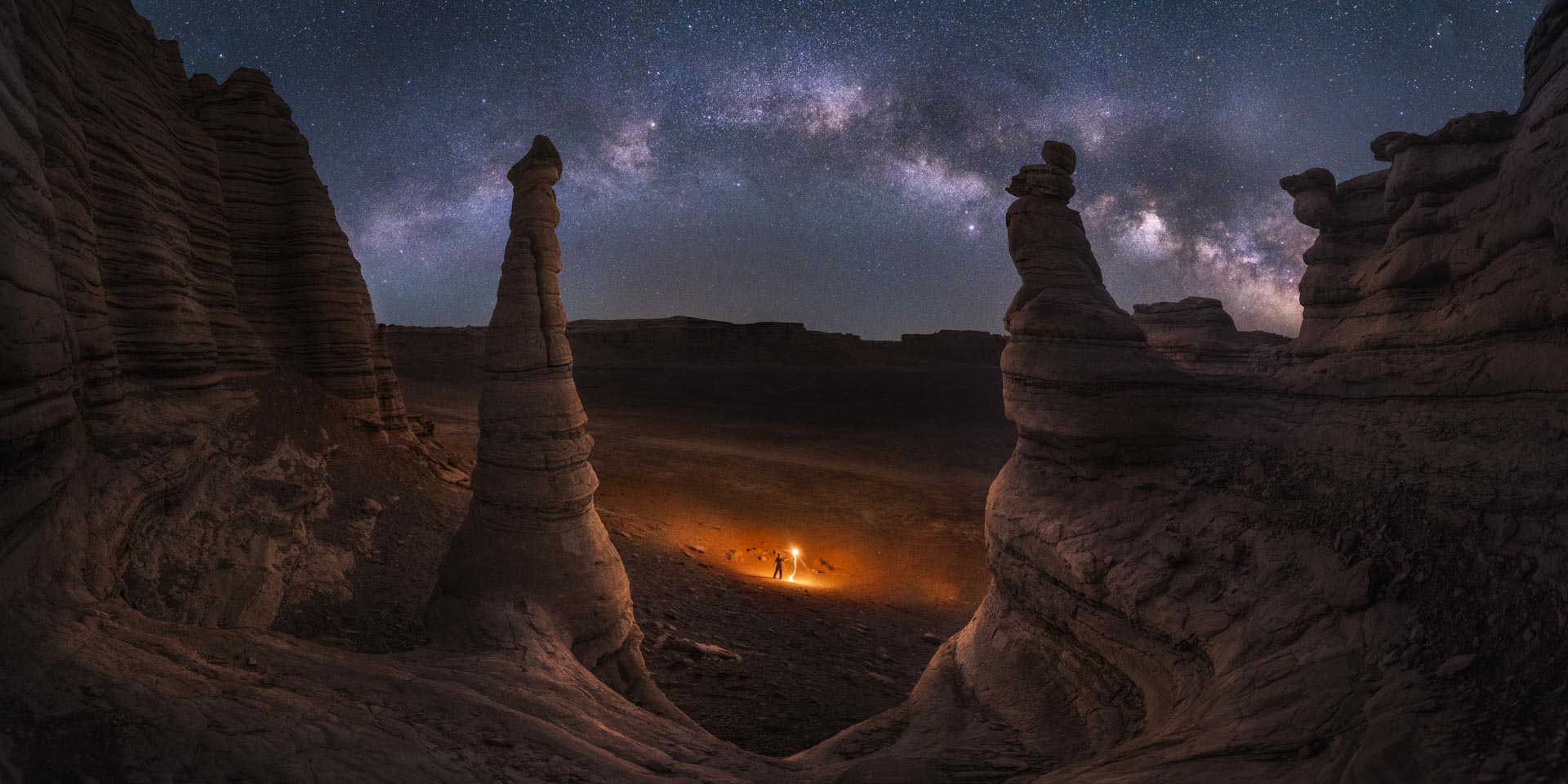Eine Canyonlandschaft bei Nacht, im Vordergrund Steinsäulen, in der Ferne eine Person mit einem licht und am Himmel die Milchstraße
