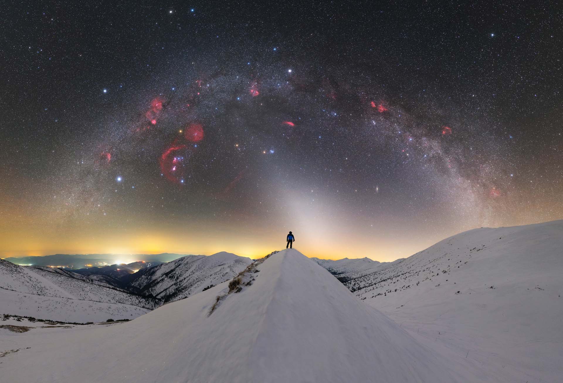 Eine Nachtaufnahme von schneebedeckten Bergen mit einer Person im Mittelpunkt auf einem Gipfel, über dem Himmel erstreckt sich die Milchstraße in einem Bogen