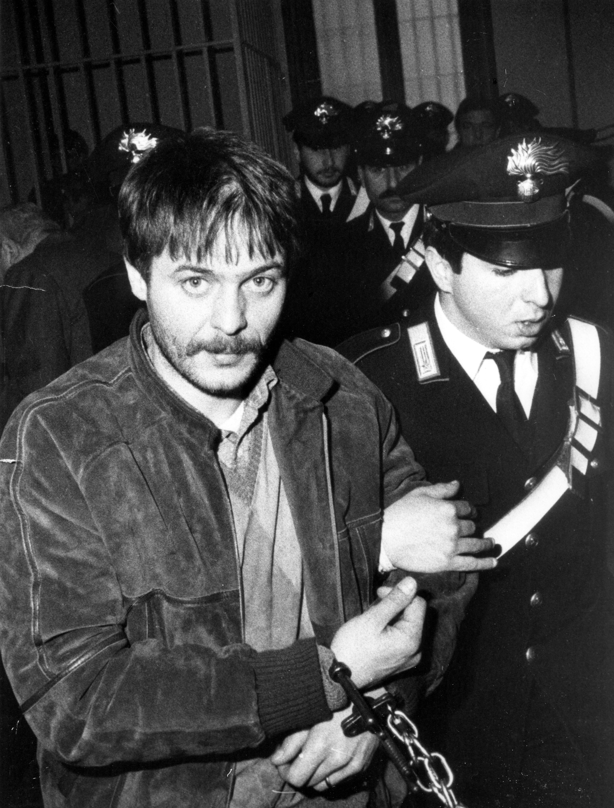 Ein junger Mann mit Schnurrbart wird in Handschellen von einem Polizisten abgeführt und schaut direkt in die Kamera