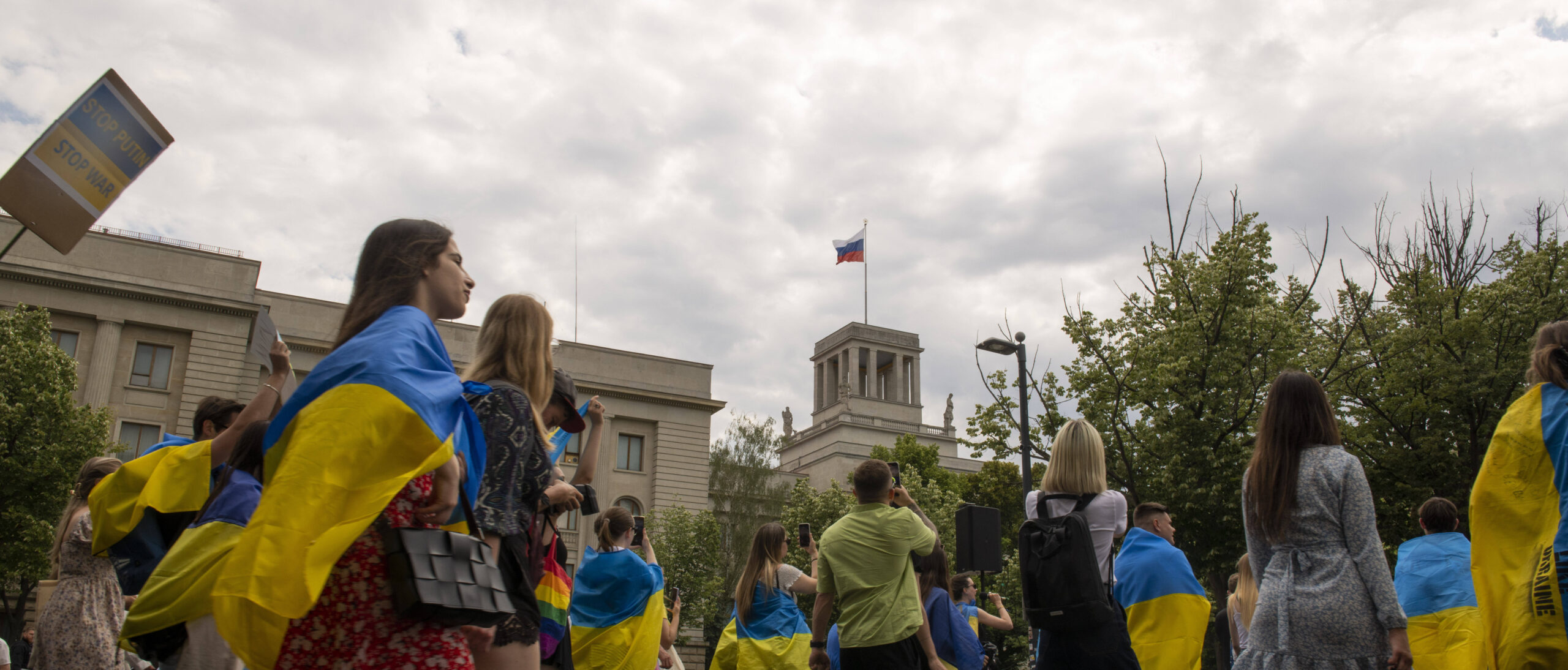 Menschen demonstrieren vor der russischen Botschaft in Berlin. Sie haben sich ukrainische Flaggen um die Schultern gebunden.