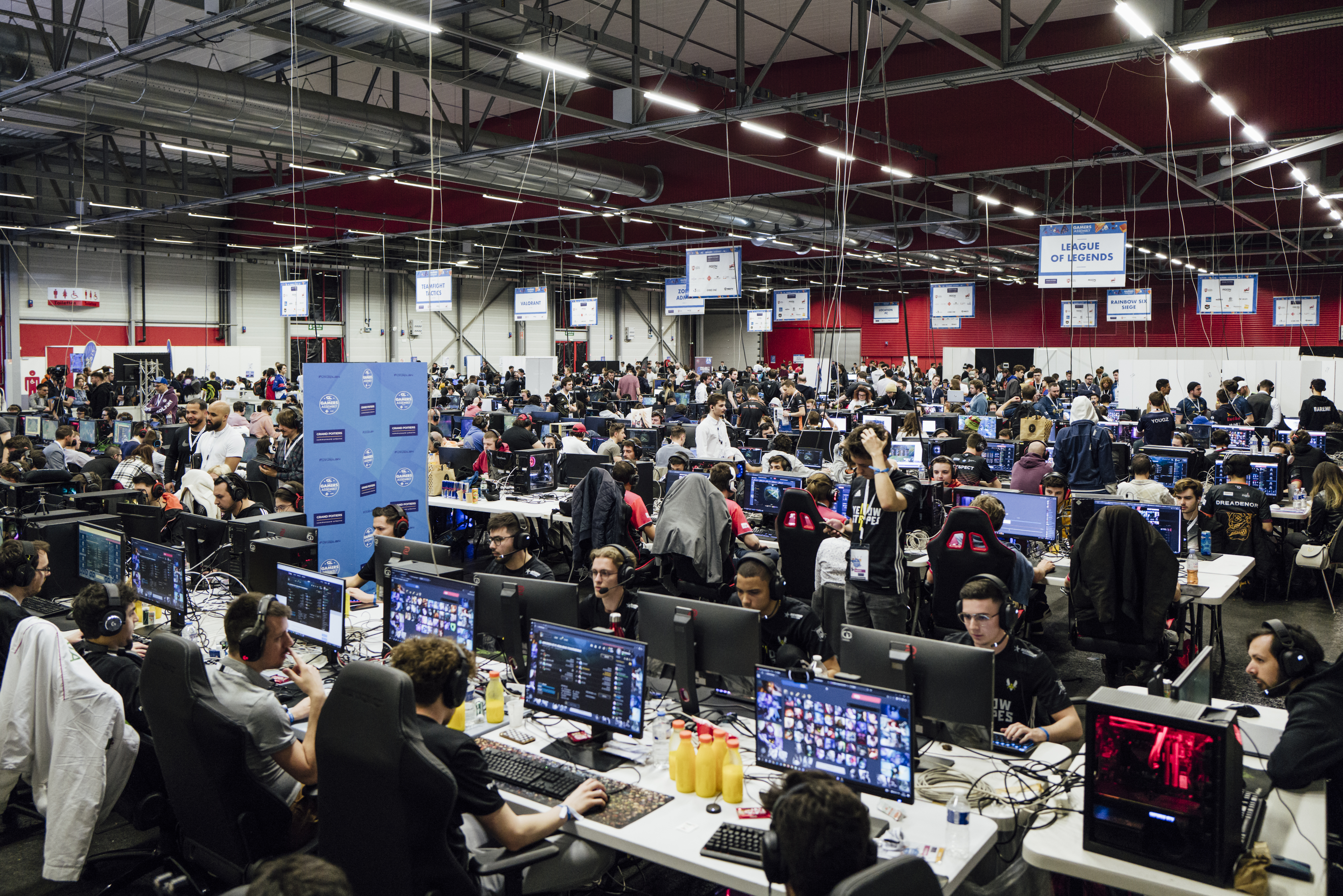 Gamers Assembly, LAN party – A giant industrial room filled with long white desks where people are sitting in front of large computer screens.
