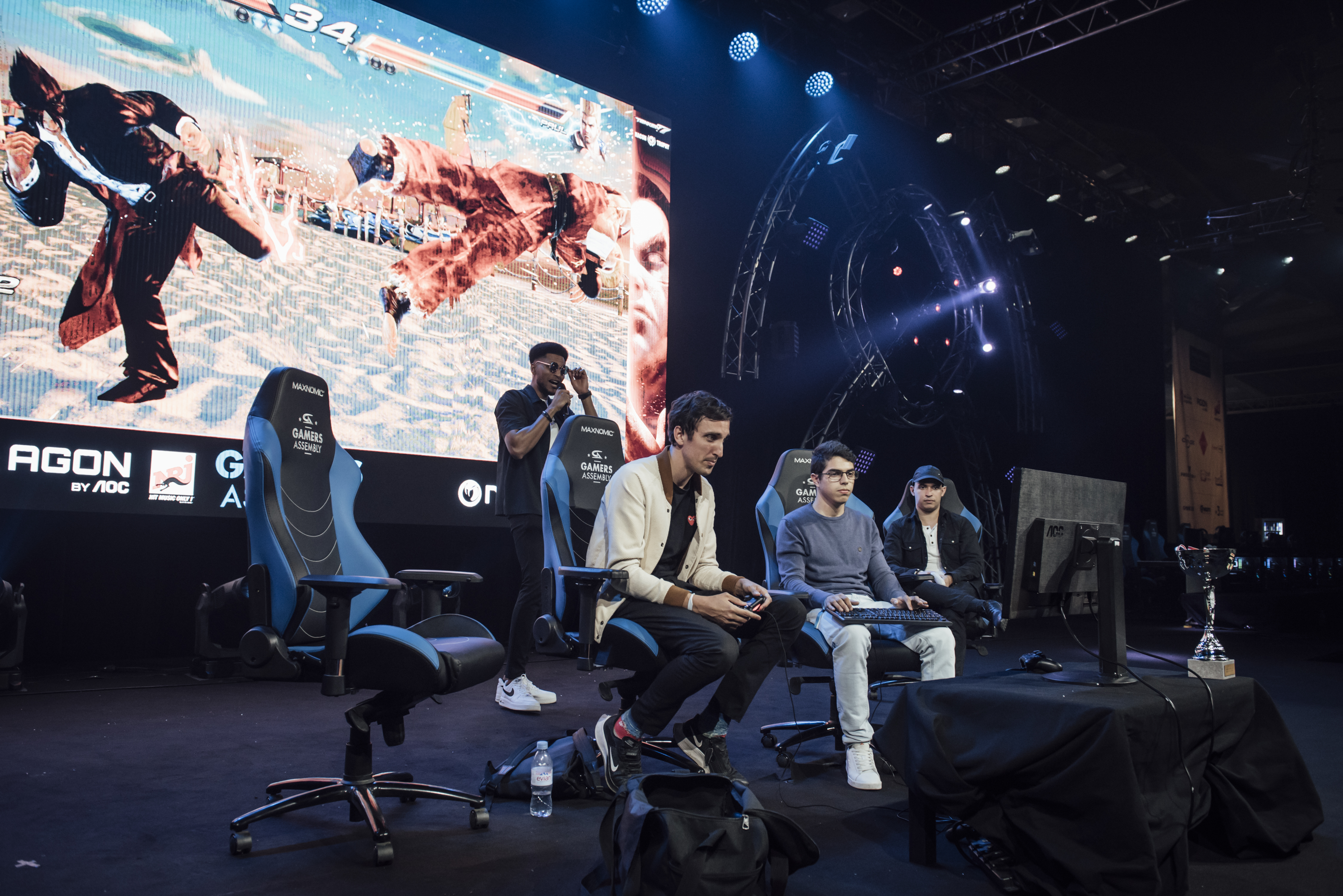 Gamers Assembly, LAN party – Two young men sitting on gaming chairs and playing tekken. In the background, a large screen is showing their fight to the public.