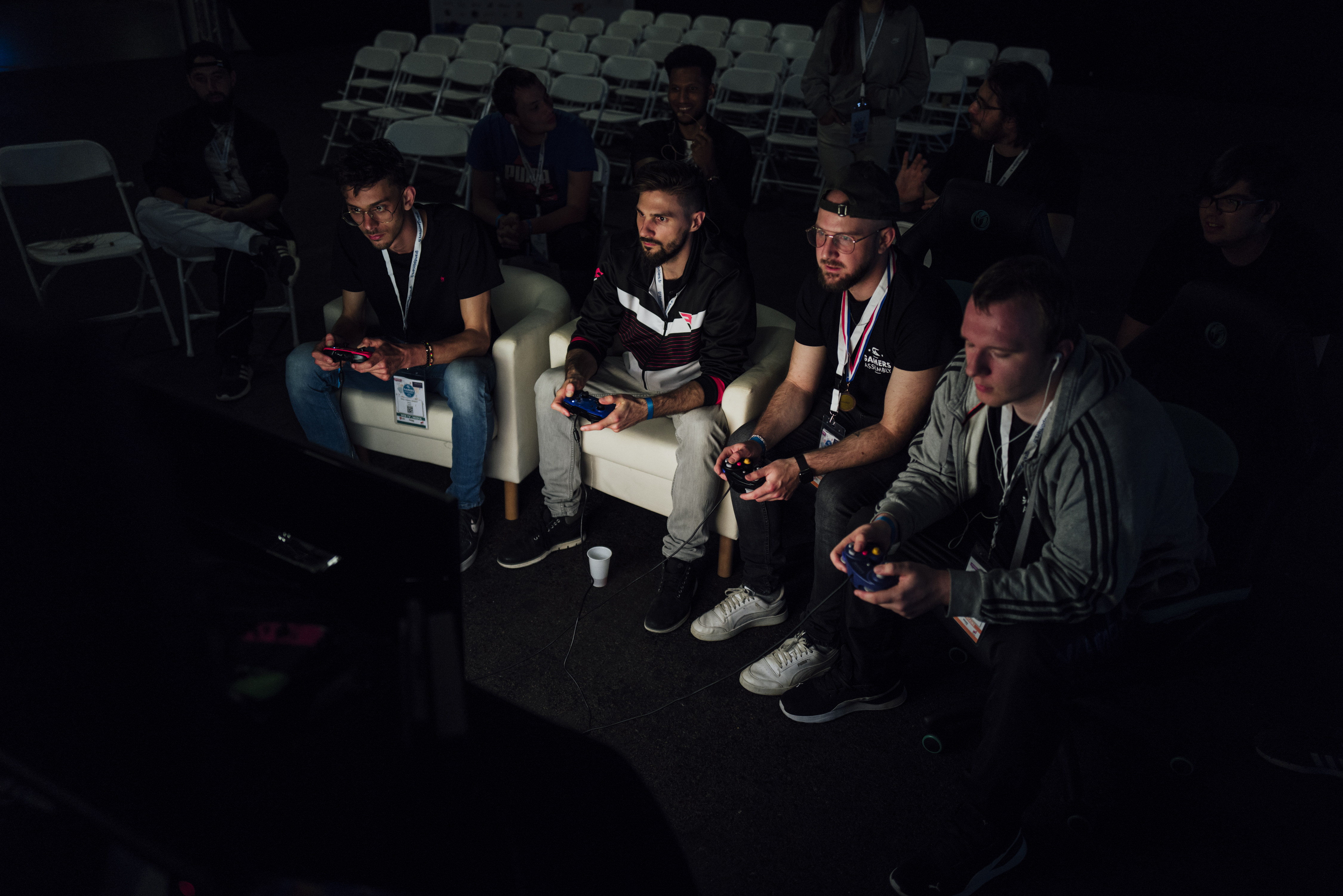 Gamers Assembly, LAN party – Four guys sitting on white chairs in an empty, dark room and playing in front of a screen.