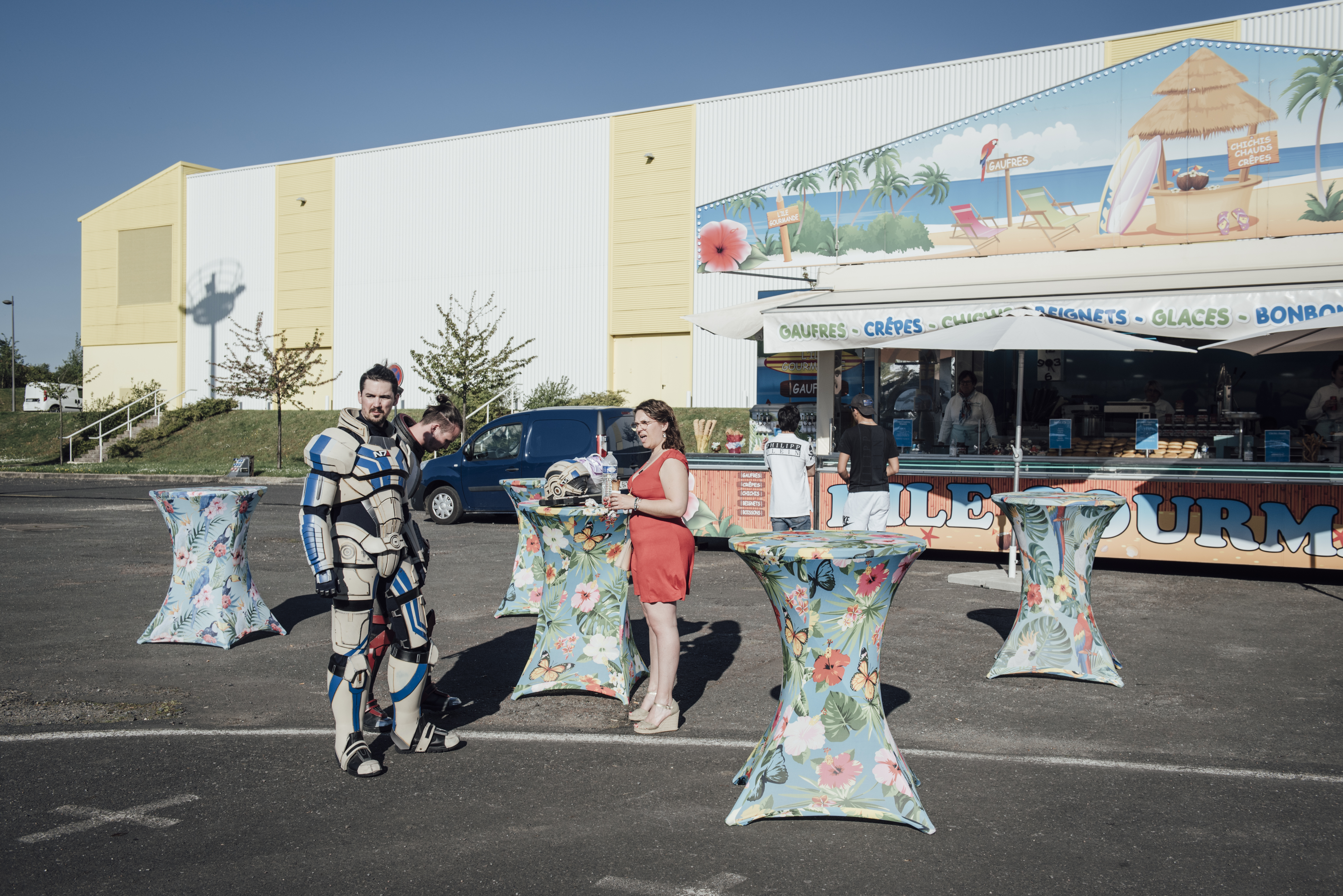 Gamers Assembly, LAN party – three people standing in front of a stall selling crepes, waffles and ice cream. A woman is wearing a short dress and heels, the two man are cosplaying in armoured suits.