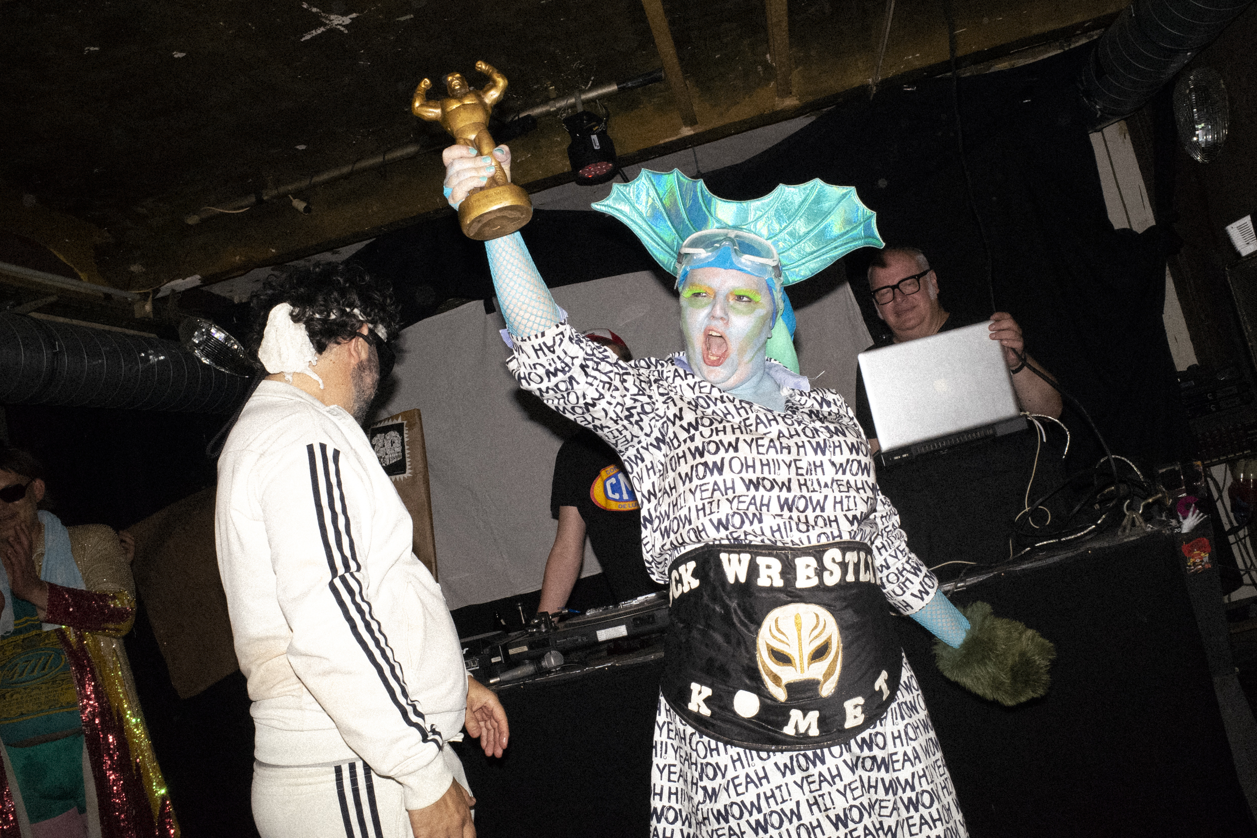 Rock & Wrestling, Hamburg – woman painted in light blue with a shiny, fin-shaped headpiece, lime green eyelashes and blue fishnet gloves, wearing a black an white onesie with writings on it and showing off her trophy and belt in front of the dj set