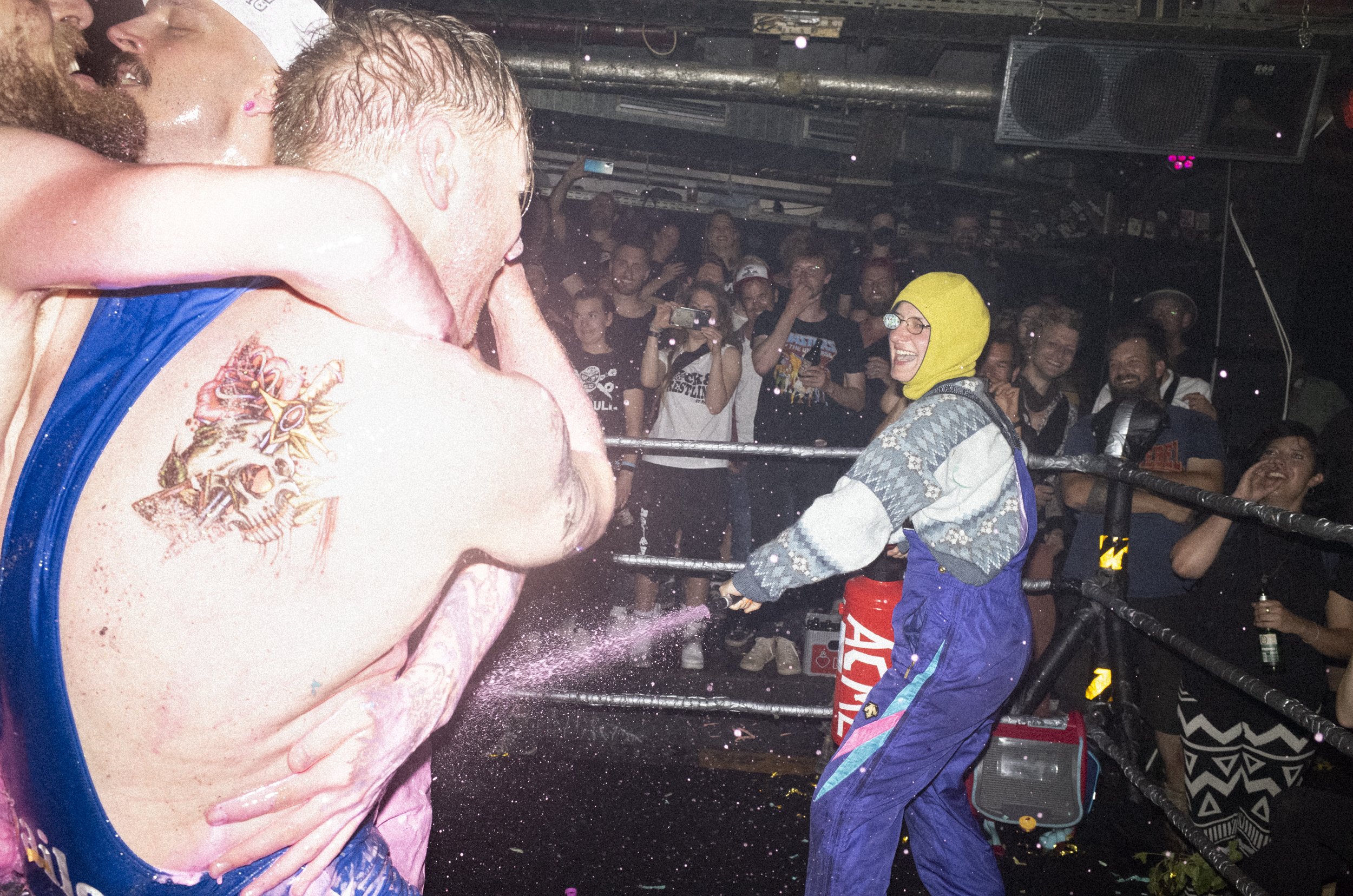 Rock & Wrestling, Hamburg – person in a purple ski suit and grey jumper spraying a group of men that locked in a sweaty embrace inside the ring as the crowd cheers on.