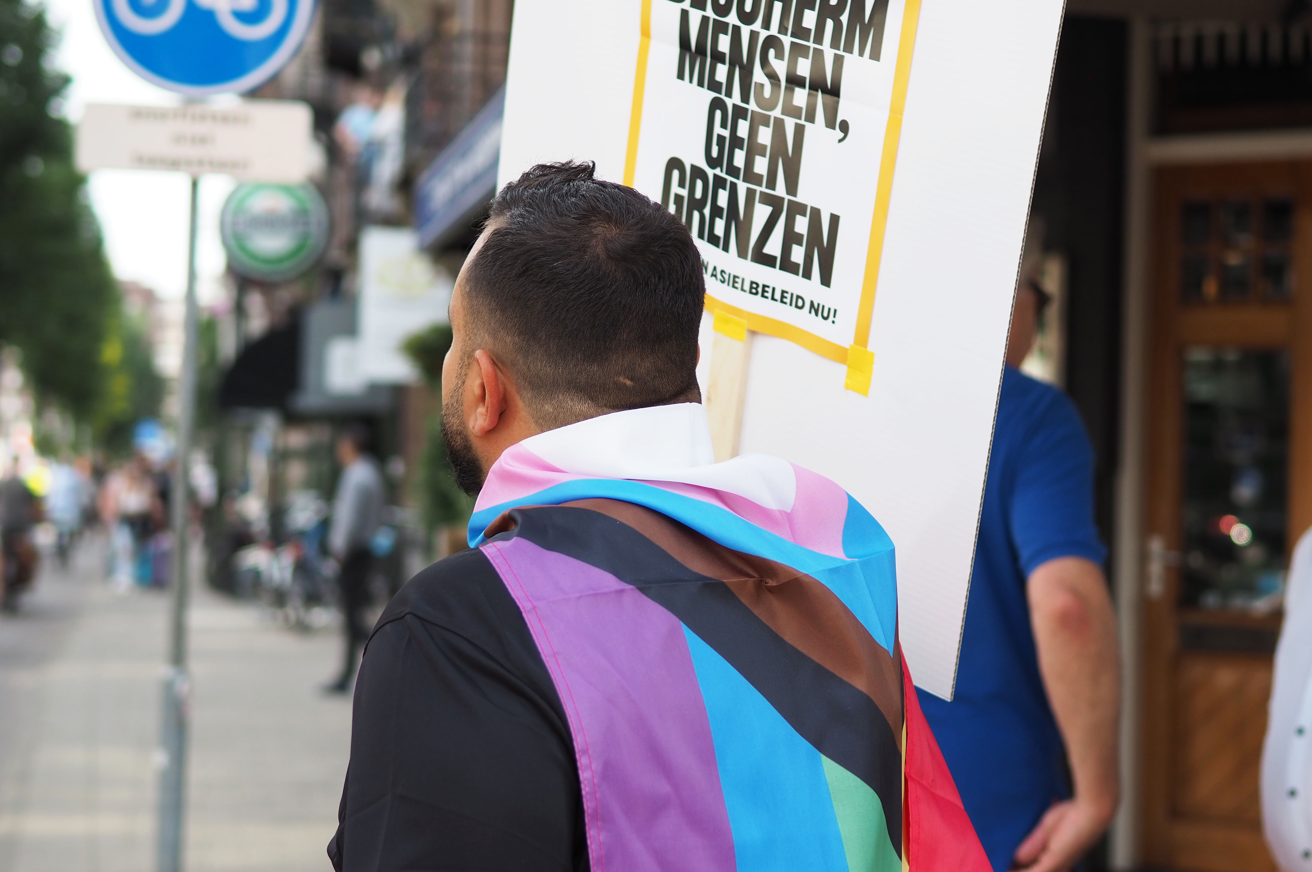 Sam – picture taken from behind of a man with short dark hair wrapped in a progressive pride flag and holding a protest sign.