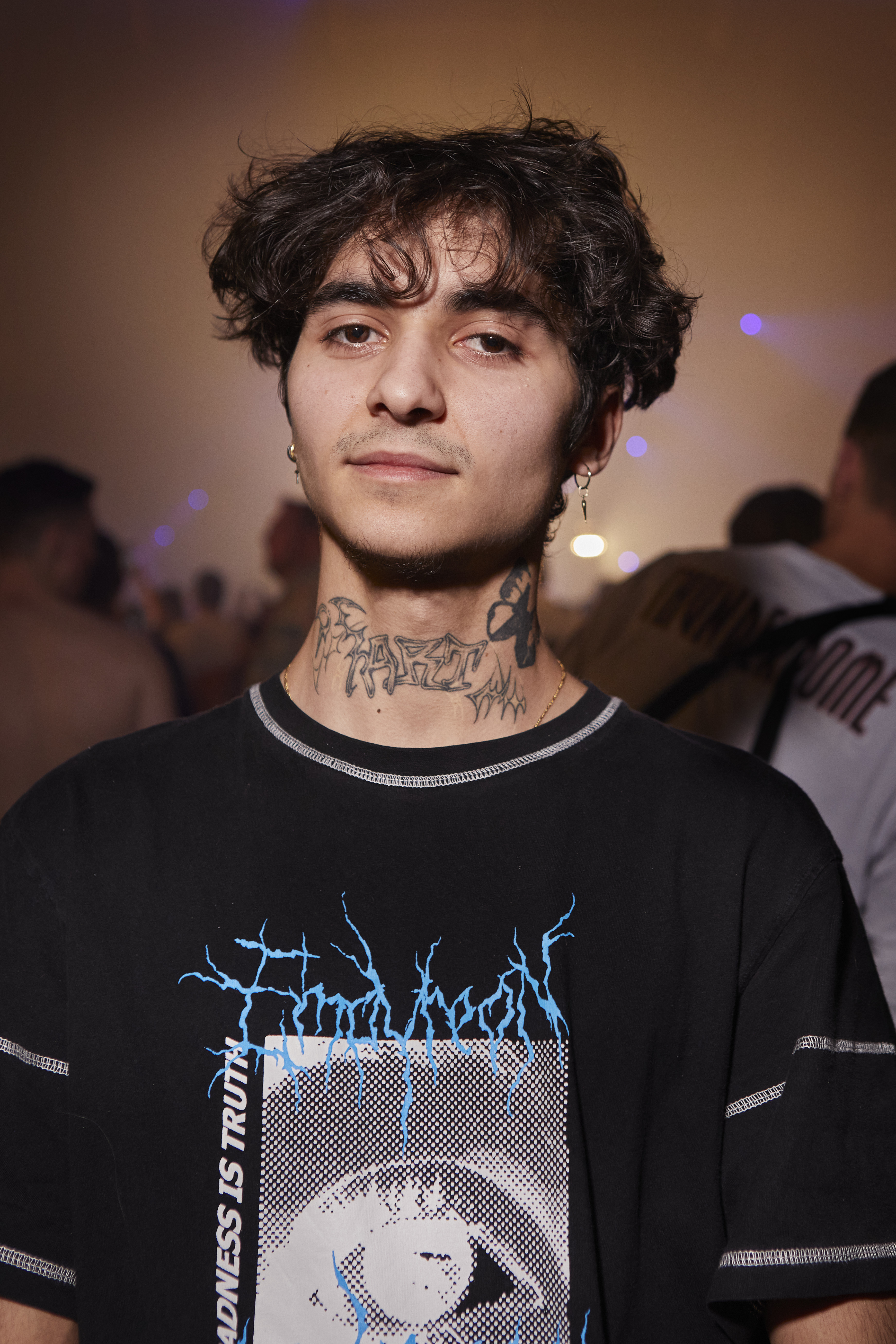 Portrait of a young man with brown curly hair and brown eyes. He has earrings and tattoos and his nick and wears a black t-shirt with blue and white patterns