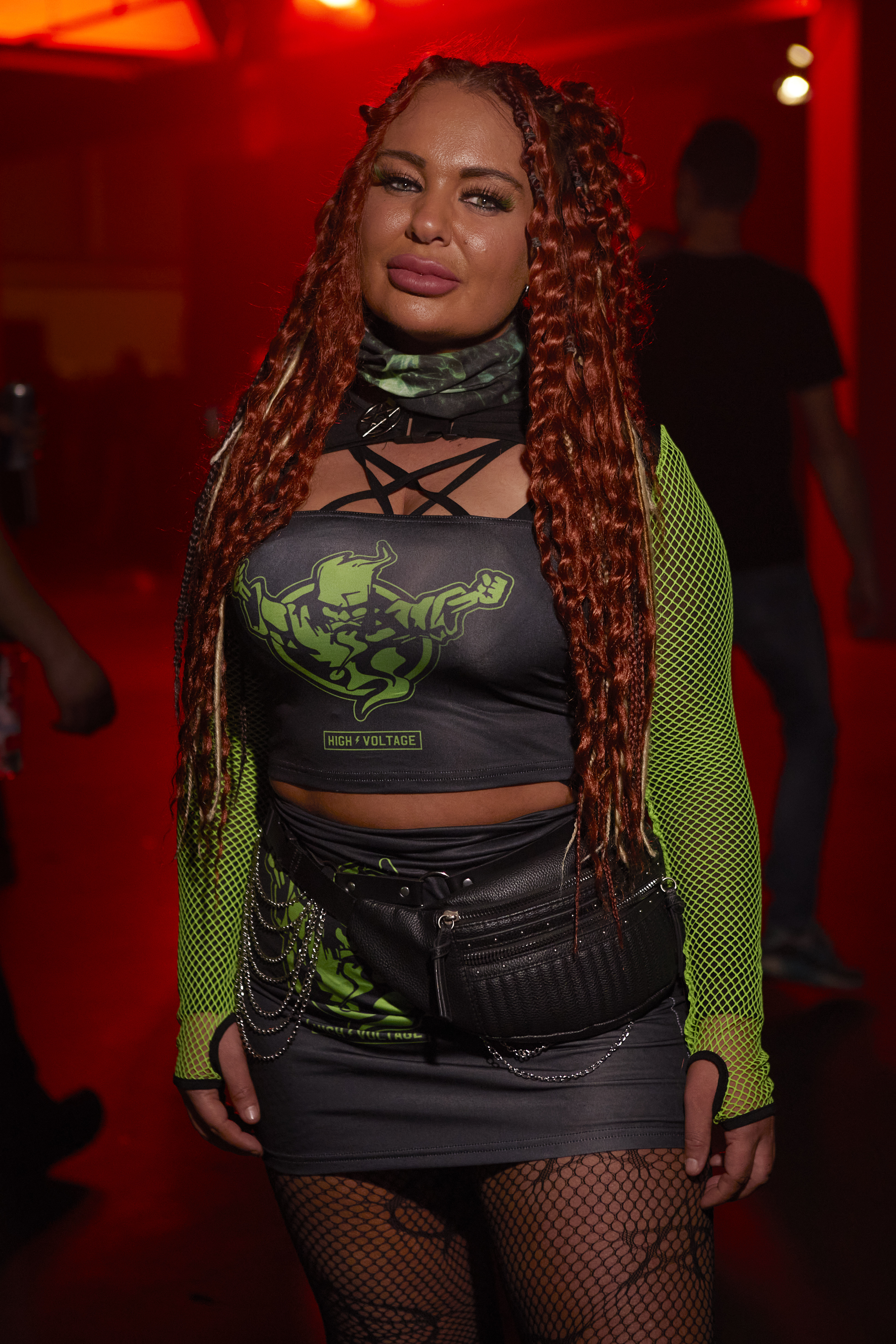 Portrait of a woman with red braids and locks. She's wearing neon green net sleeves, and a black and green party outfit with the Thunderdome logo