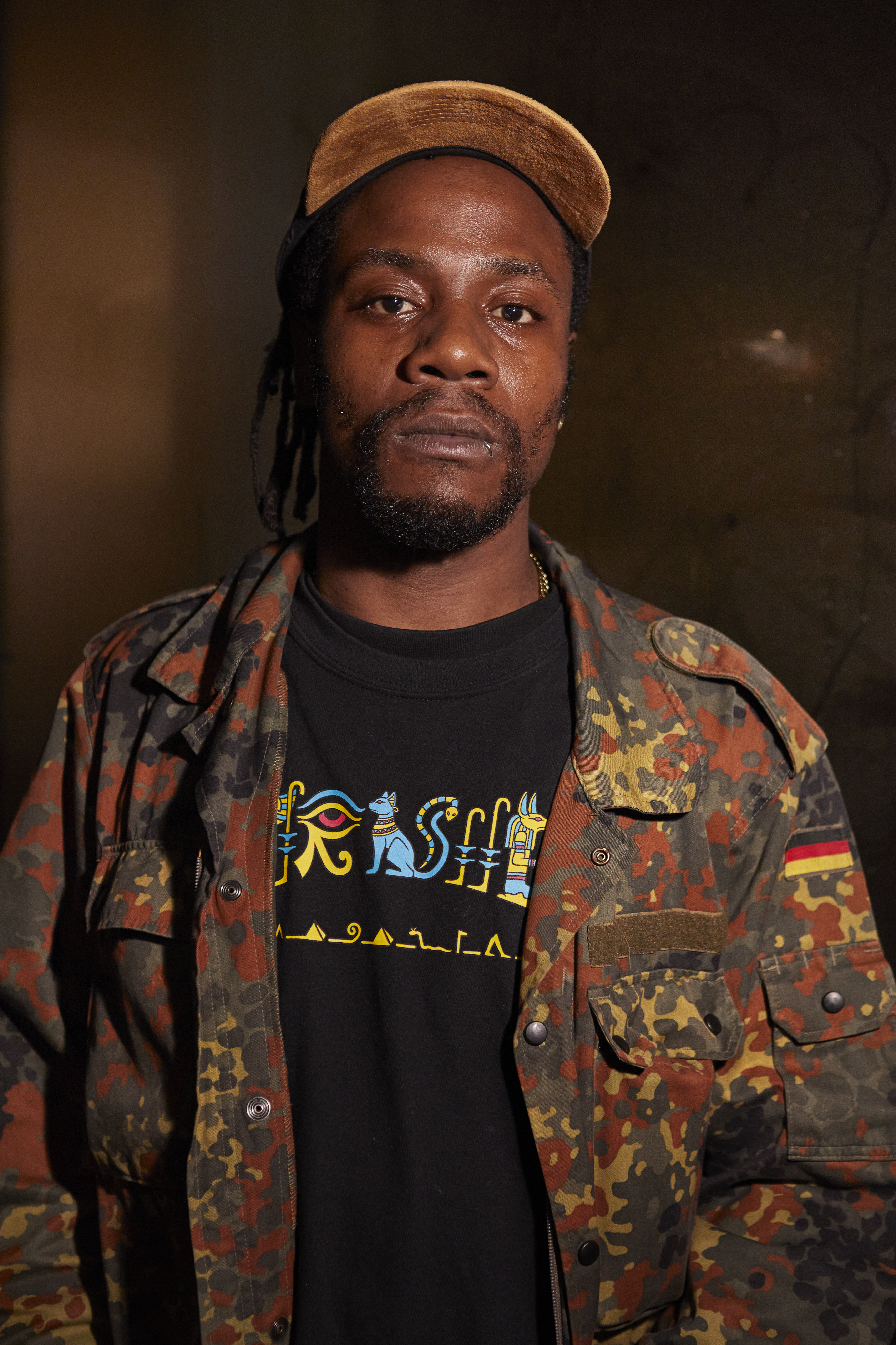 Portrait of a man with black deadlocks and a beard, wearing a cameo jacket, on a black t-shirt with blue and yellow hieroglyphs and a cap
