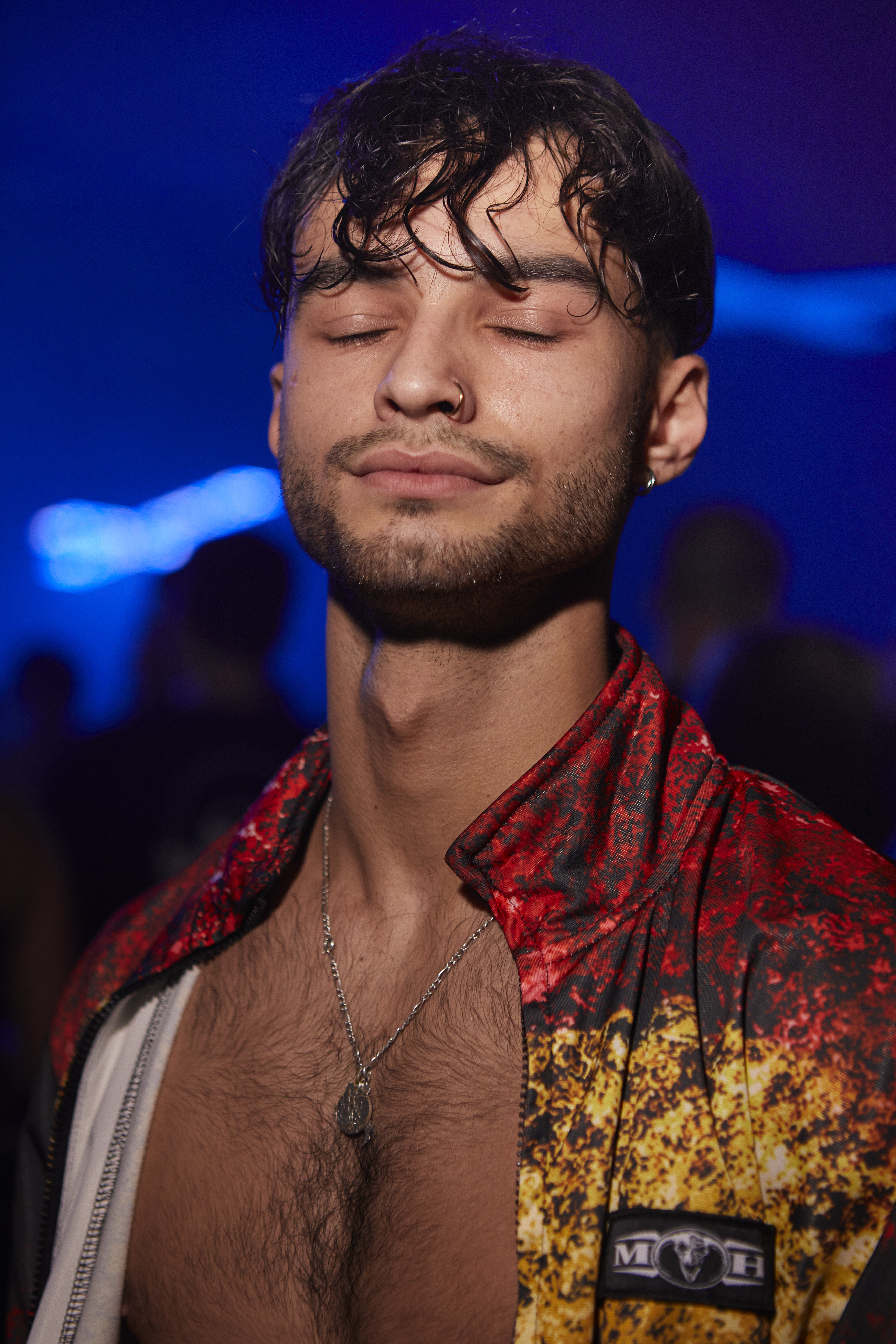 Portrait of a topless man with dark hair and a nose piercing, he's wearing an open red and yellow jacket, his hair is wet and he's closing his eyes