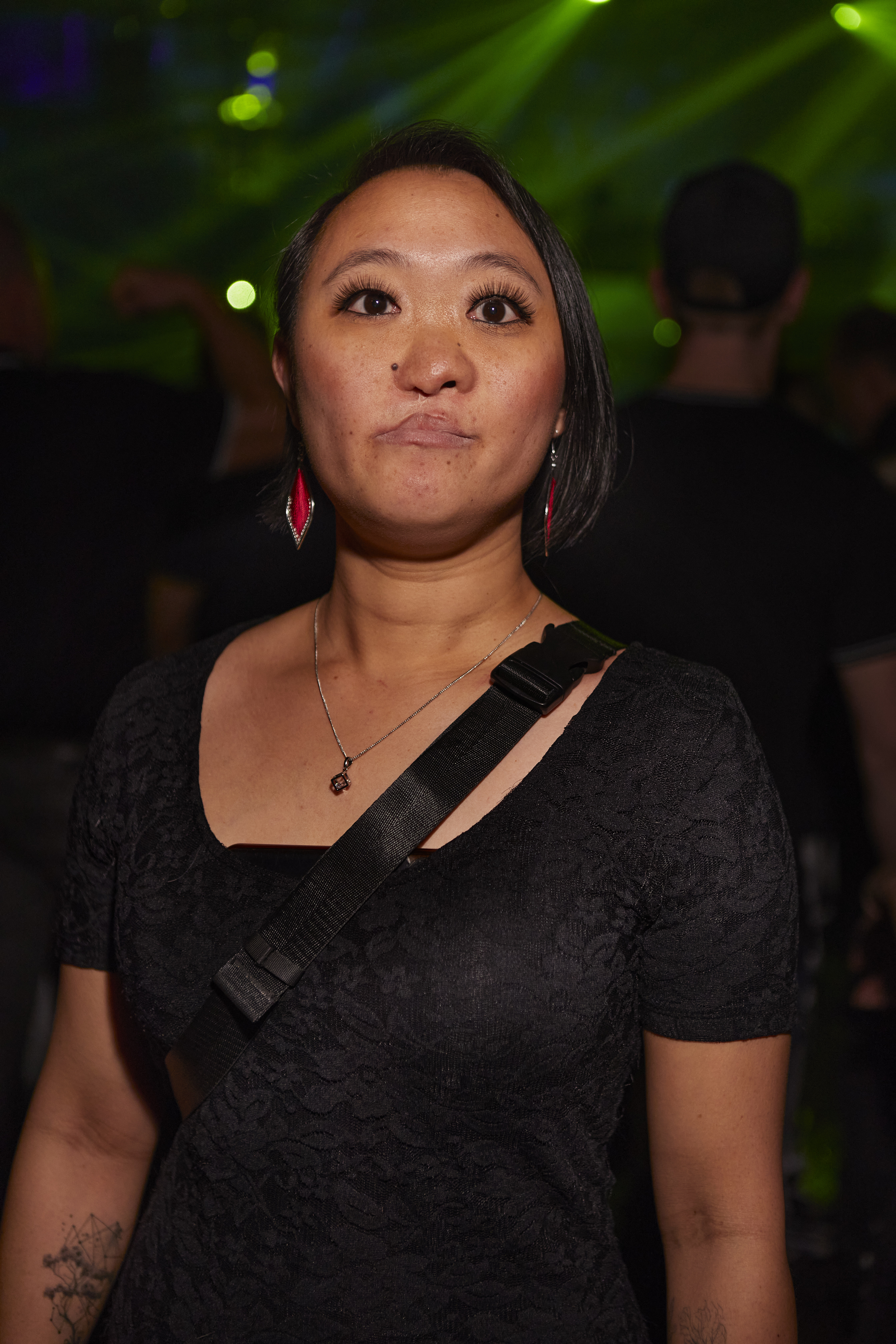Portrait of a woman with black straight hair wearing a black tshirt and red earrings. She has a tattoo on her right arm