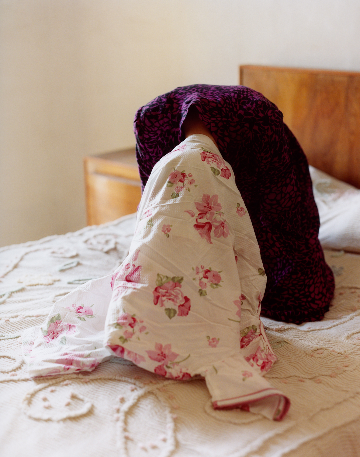 A person sitting on a bed hunches over with a floral cloth covering their entire head.