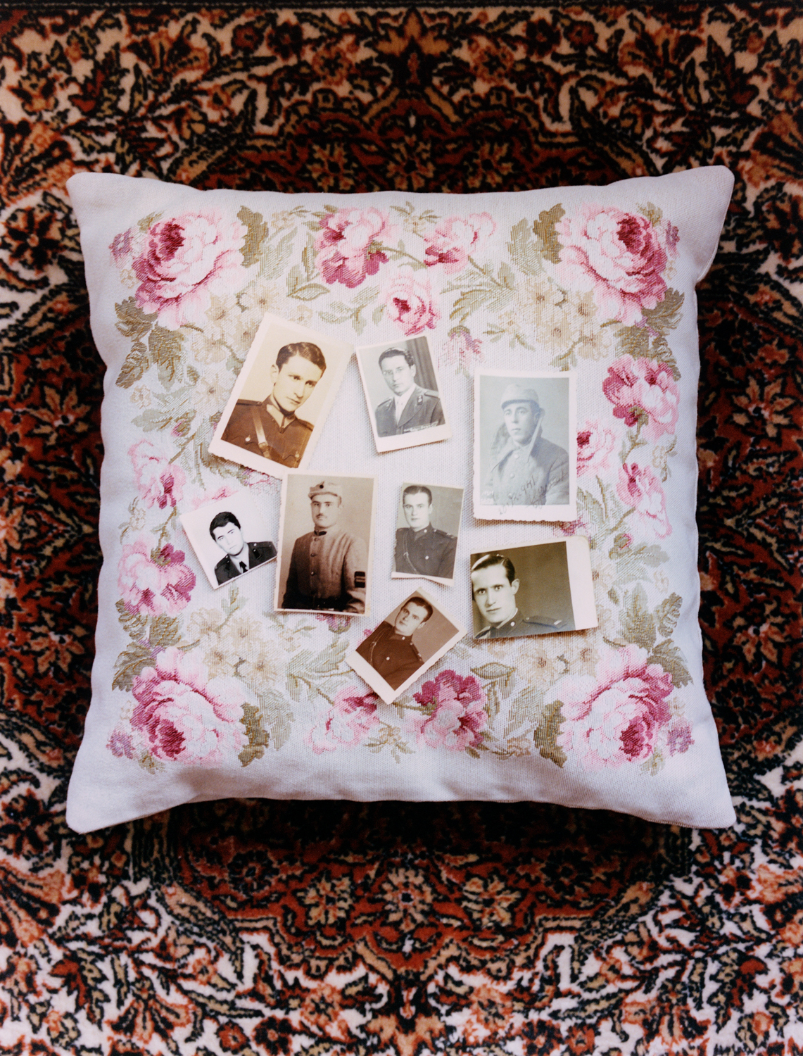A collection of photographs of uniformed men sit on top of a floral throw pillow, the pillow is on top of a rug.