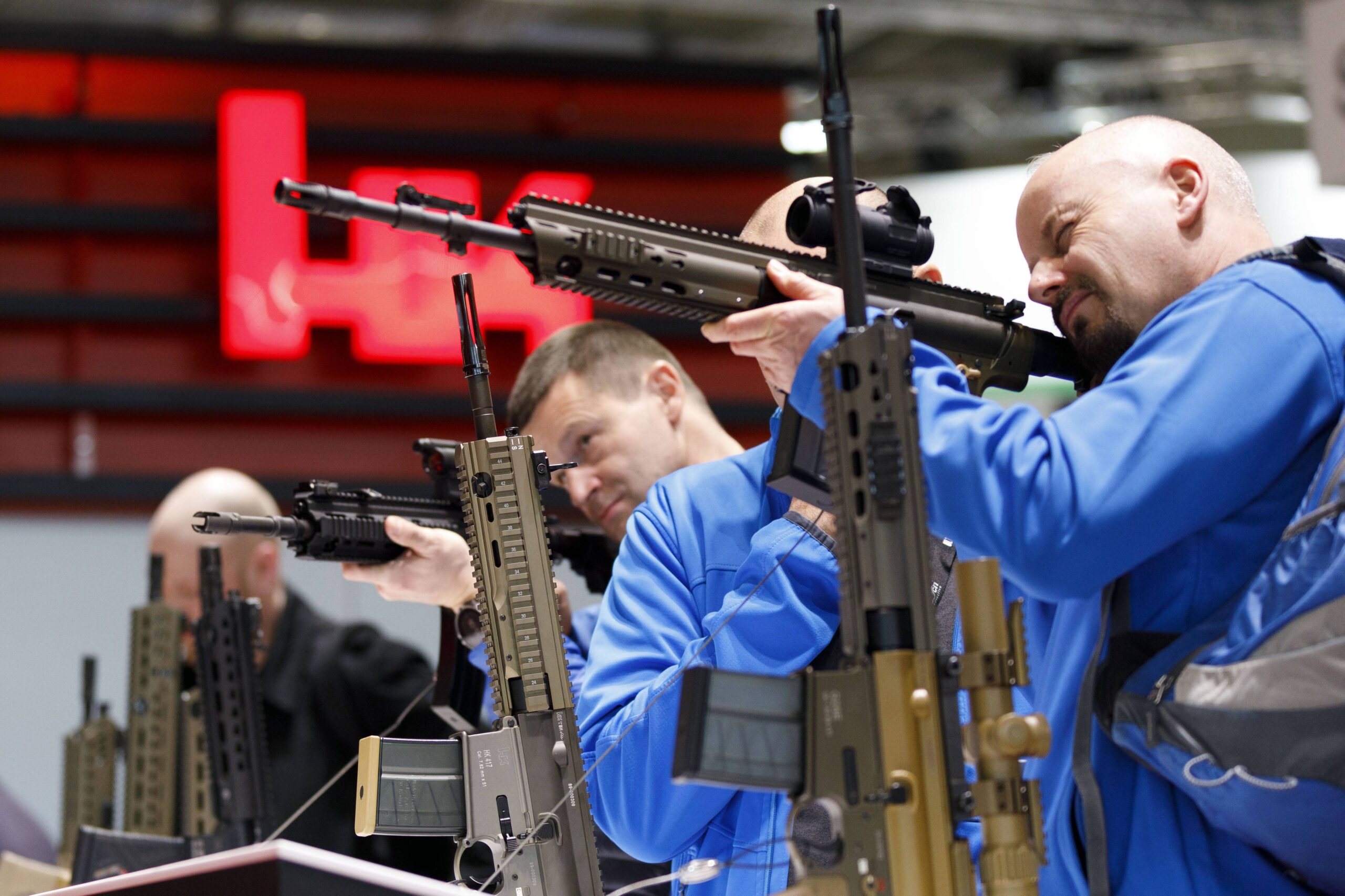Ein glatzköpfiger Mann in blauer Jacke hat ein Sturmgewehr an seine Schulter gelegt, kneift ein Auge zusammen und zielt.