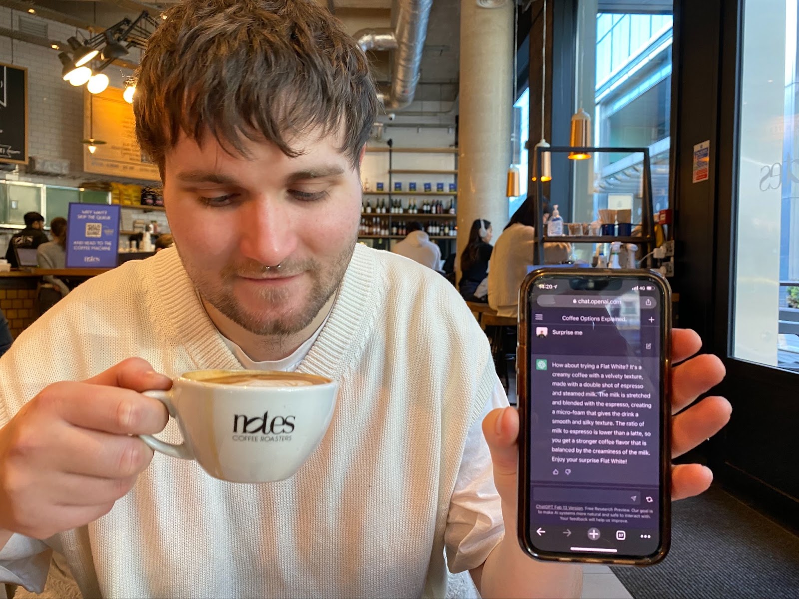 A man drinking coffee and holding up a ChatGPT screen on his phone