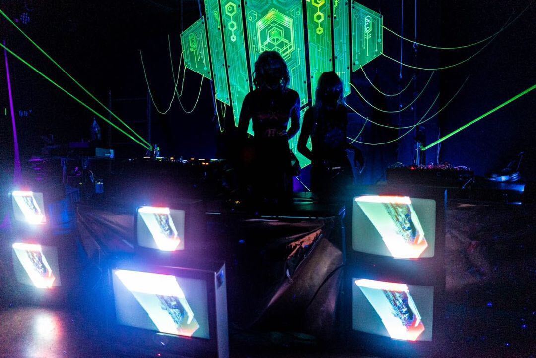 The silhouettes of two people standing in front of a green laser cut sculpture and surrounded by a wall of TV monitors