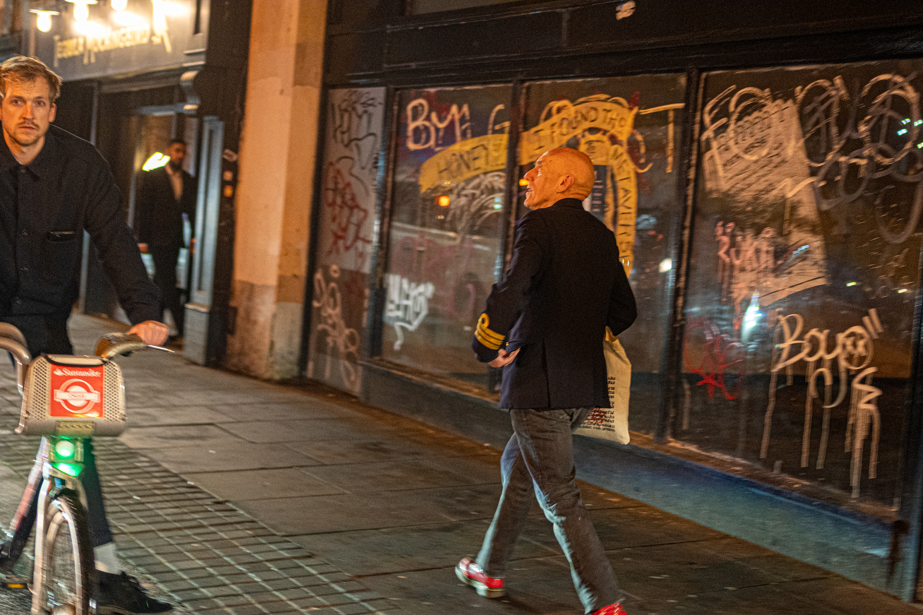 Roger walks down a dark street lit by orange streetlights, behind him are graffitied windows, next to him is a young man riding a bike.