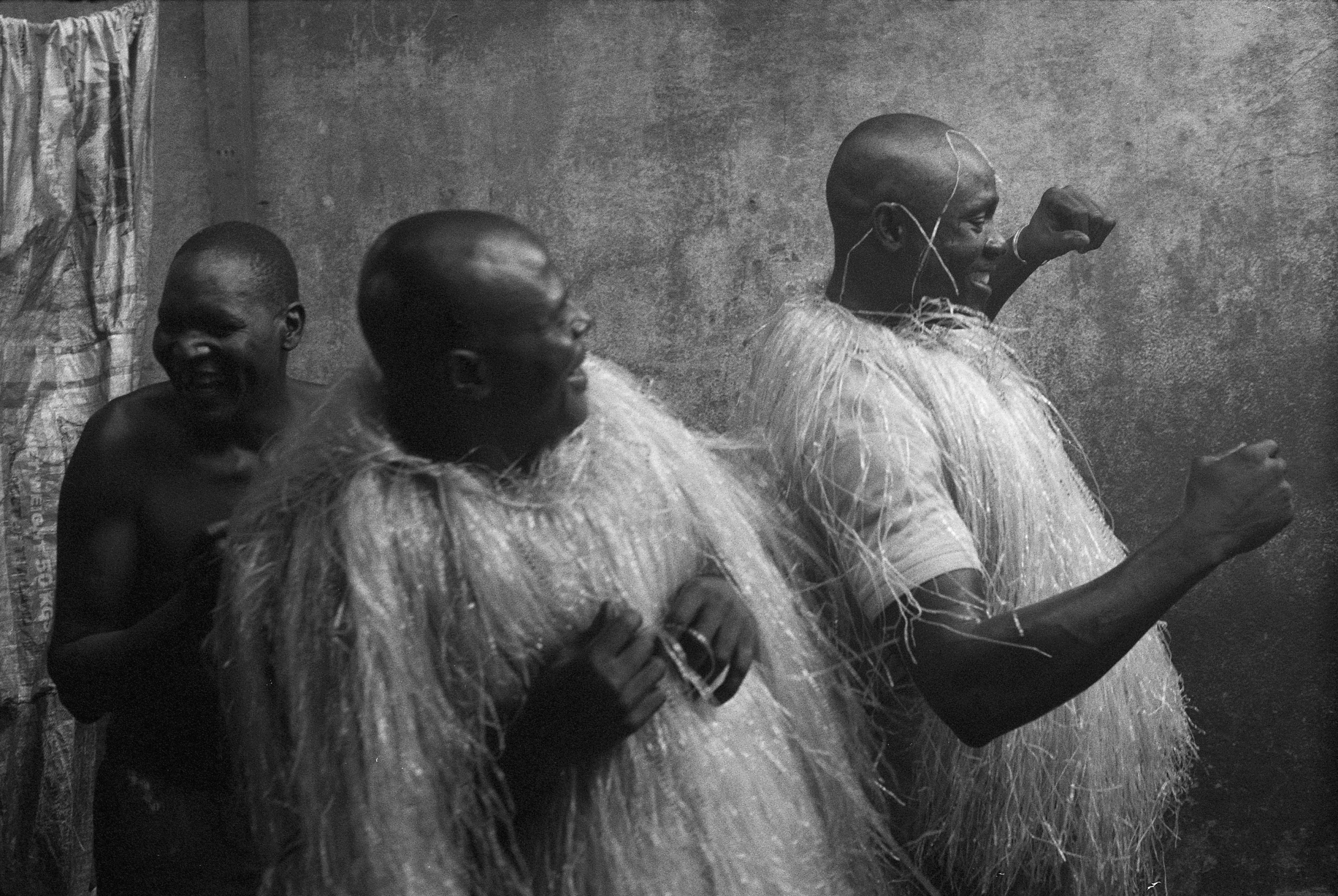 Schwarz-weiß-Foto von drei Männern, die lachend tanzen, zwei tragen dabei Kostüme, die an traditionelle afrikanische Gewänder erinnern