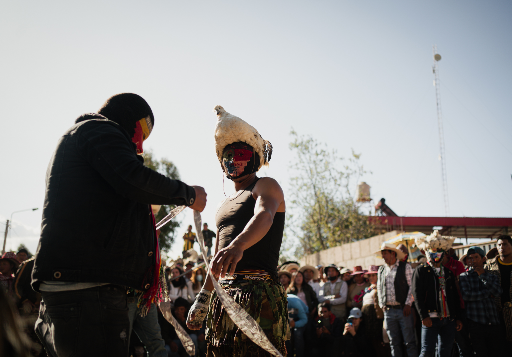 Takanakuy, Peru – man wearing mask and biker jacket, wrapping the hands of another man in a tank top, kaki pants, a decorated ski mask and a bird-hat. Behind them, the crowd