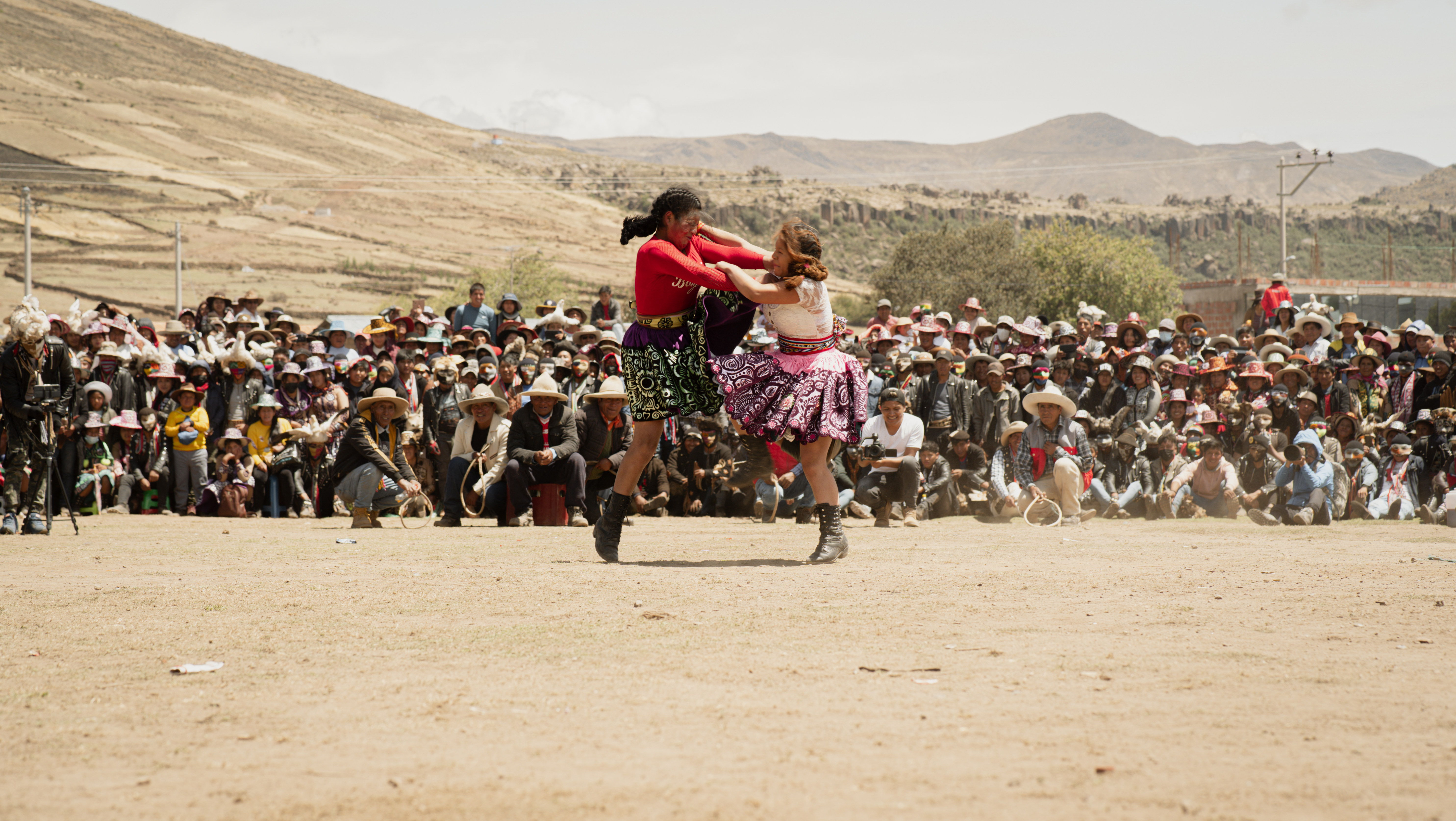 Takanakuy, Peru – two women caught mid-fight up in the air, holding on to each other's neck while kicking with the same leg. The one on the left is wearing a purple skirt with green embroidery and a red shirt. The one on the right a pink skirt with purple embroidery and a white shirt.