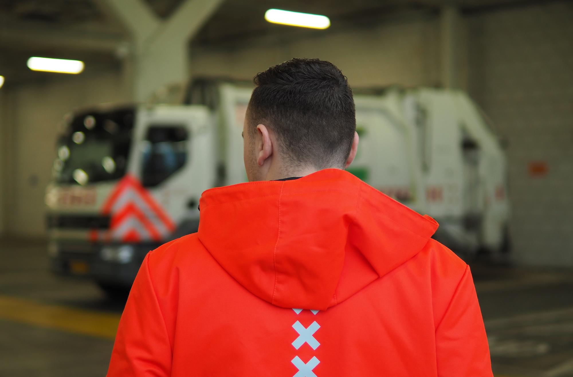 garbage collectors - portrait of a man wearing an orange uniform with reflective crosses, he's giving his back to the camera and there's a garbage truck in the background