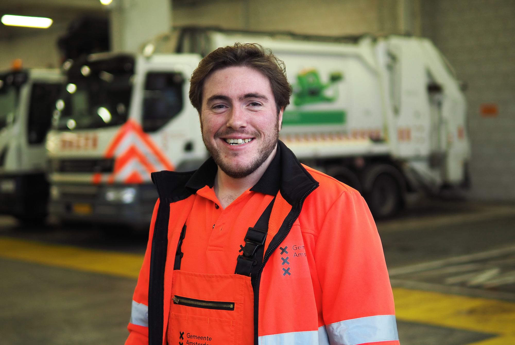 garbage collectors - portrait of a man with brown hair smiling at the camera, wearing an orange uniform with reflective bands, and there's a garbage truck in the background