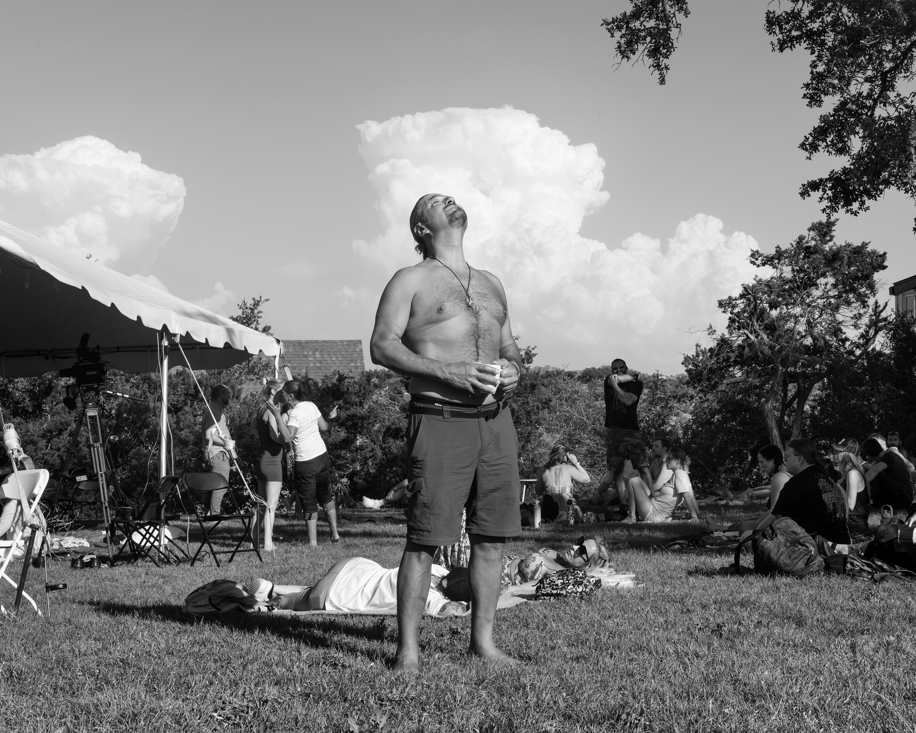 Een shirtloze man met gesloten ogen heft zijn hoofd de lucht in, achter hem zitten mensen in het gras met elkaar te praten, achter hem is een pluizige wolk te zien.