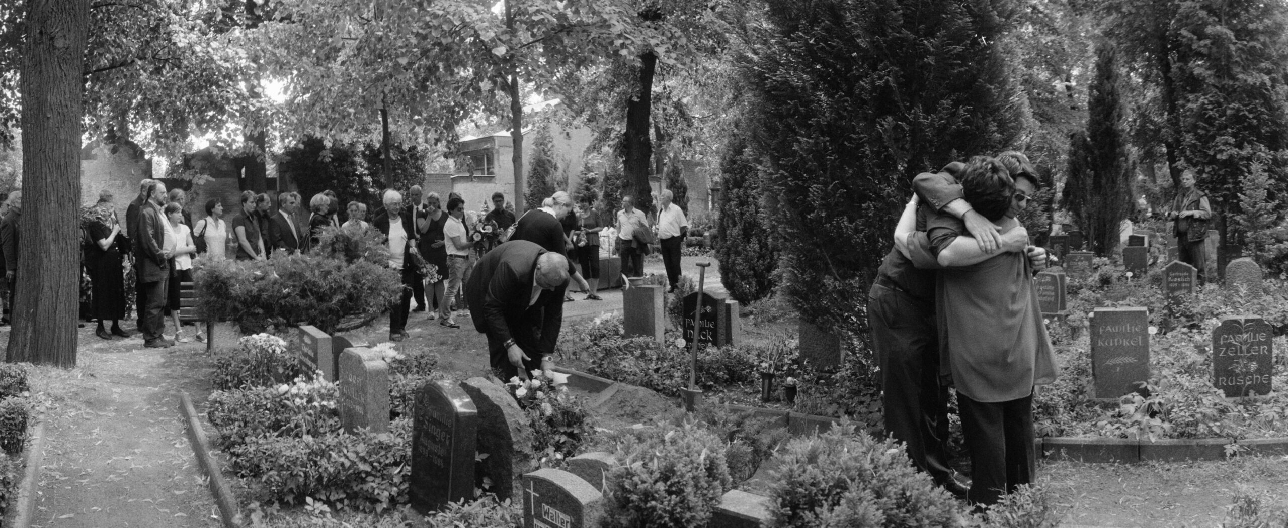Zwei Menschen umarmen sich vor einem Grab auf einem Friedhof. Im Hintergrund stehen viele Menschen und gucken zu einem anderen Grab.