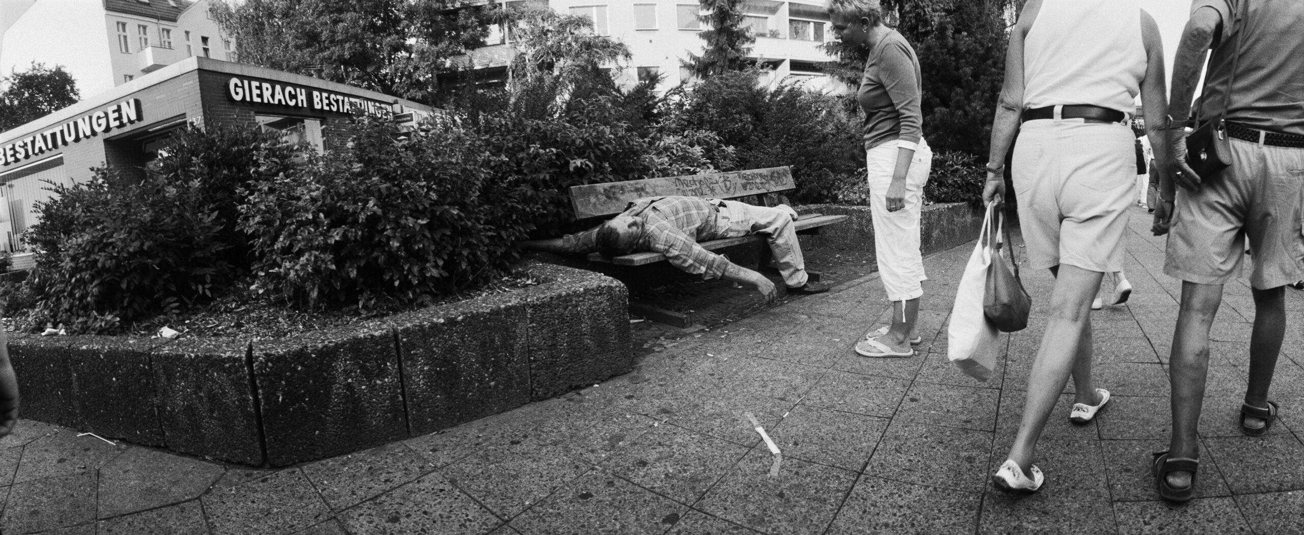 Ein Mann liegt schlafend auf einer Parkbank. Eine Frau sieht auf ihn herunter.
