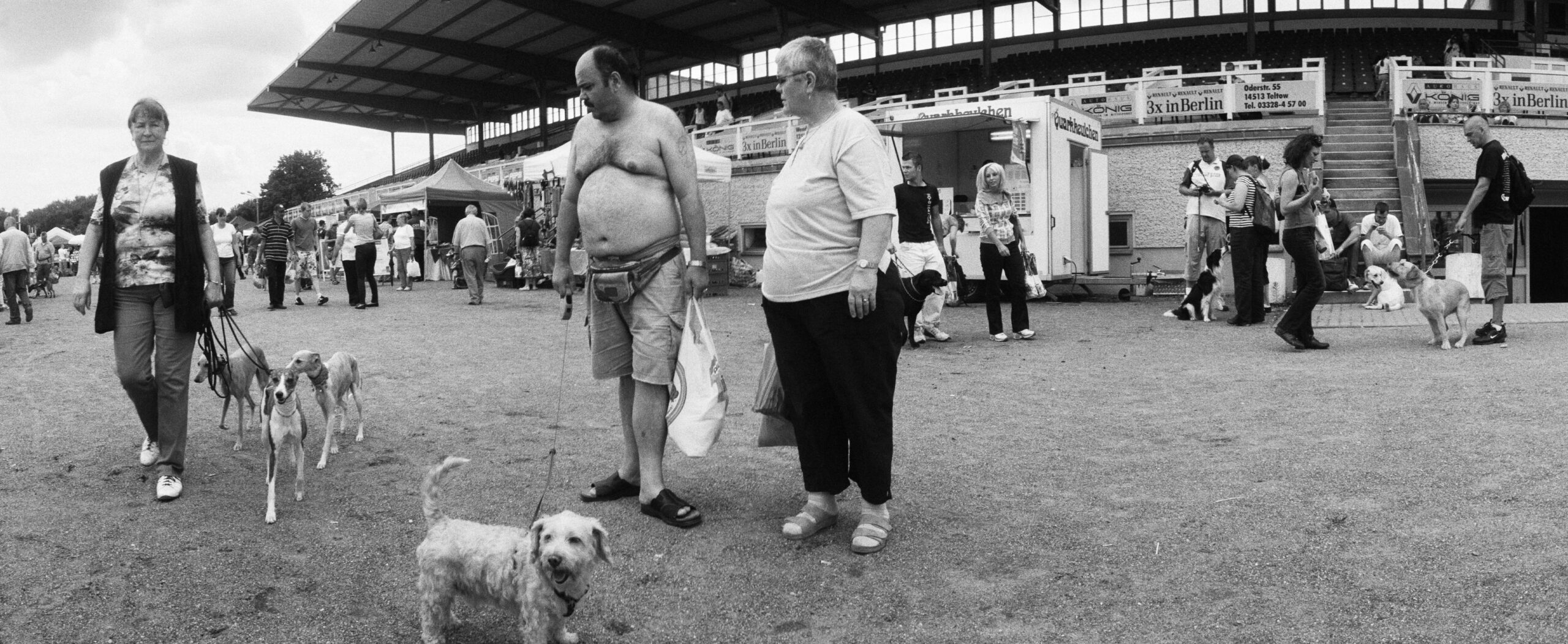 Ein Mann mit einem dicken Bauch hält einen weißen Hund an der Leine. Eine Frau mit drei anderen Hunden läuft an ihm vorbei.