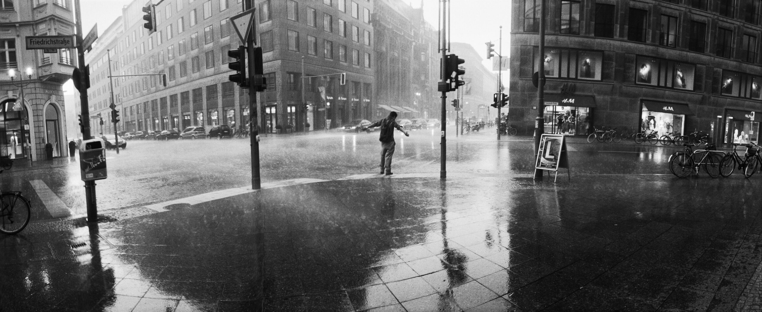 Ein Mann steht an einer großen, menschenleeren Kreuzung. Er ist der einzige auf der Kreuzung und breitet seine Arme im Regen aus.
