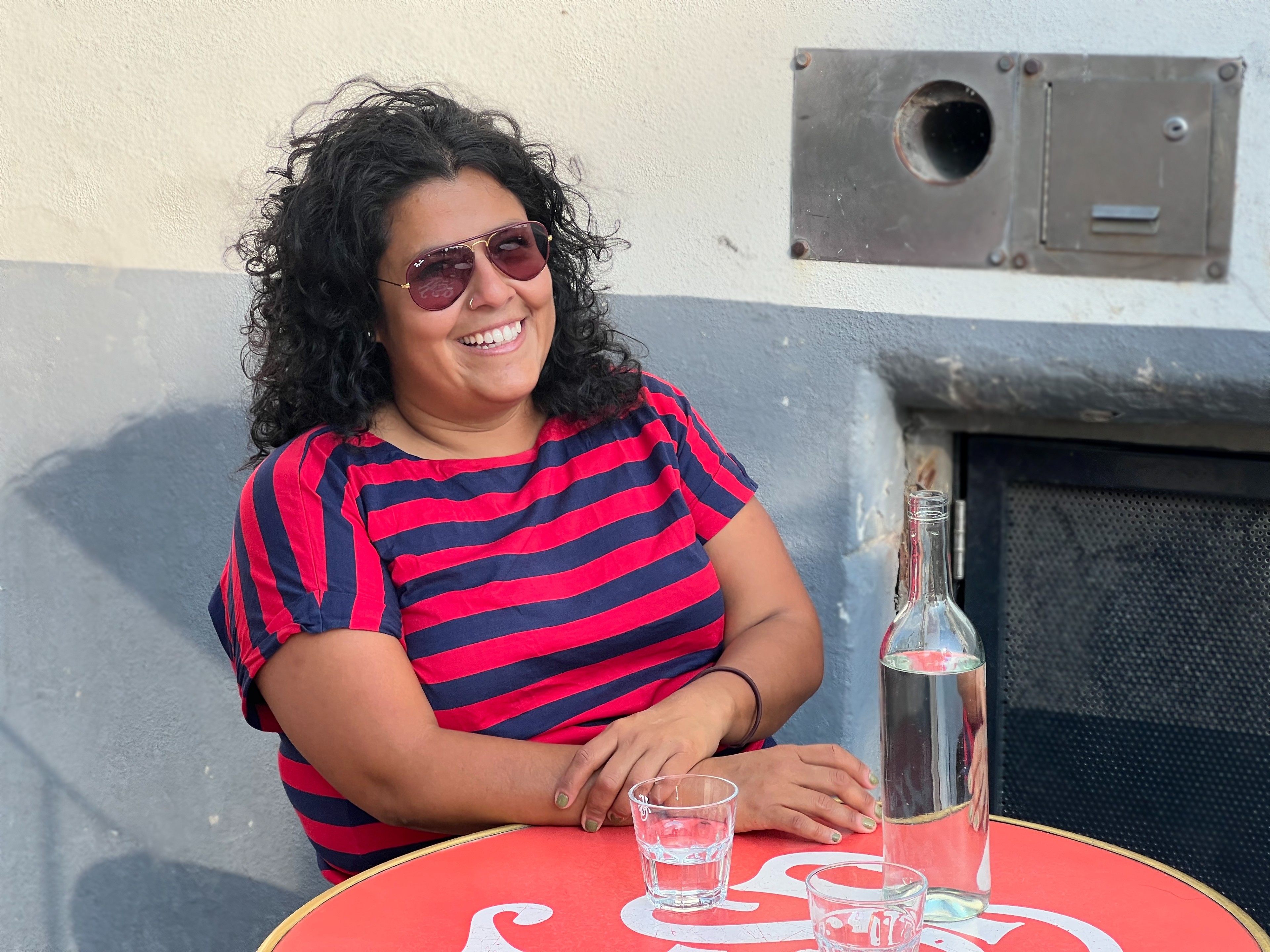 Unwanted babies, Karina – Woman with black curly hair, a blue and red striped t-shirt and aviators, sitting at a table outside in front of a bottle of water and two glasses.