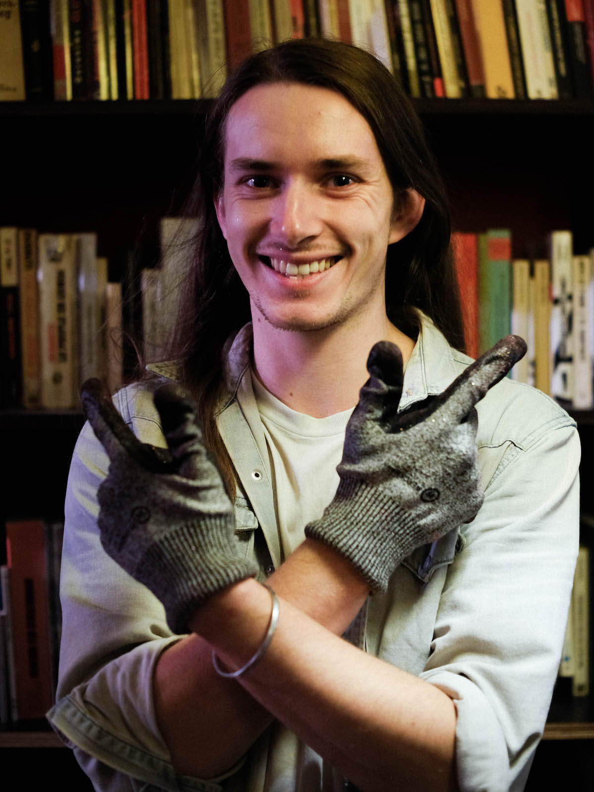 Samuel Flambard, Otoko Contraception – young man with long brown hair and brown eyes, wearing light denim shirt and utility gloves, posing with his arms crossed in front of his bodies and doing finger guns and smiling.