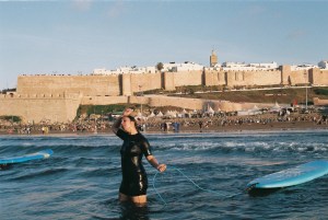 Alaeï steht im Wasser und zieht ein blaues Surfbrett hinter sich her. In dem Artikel geht es um marokkanische Frauen, die surfen.