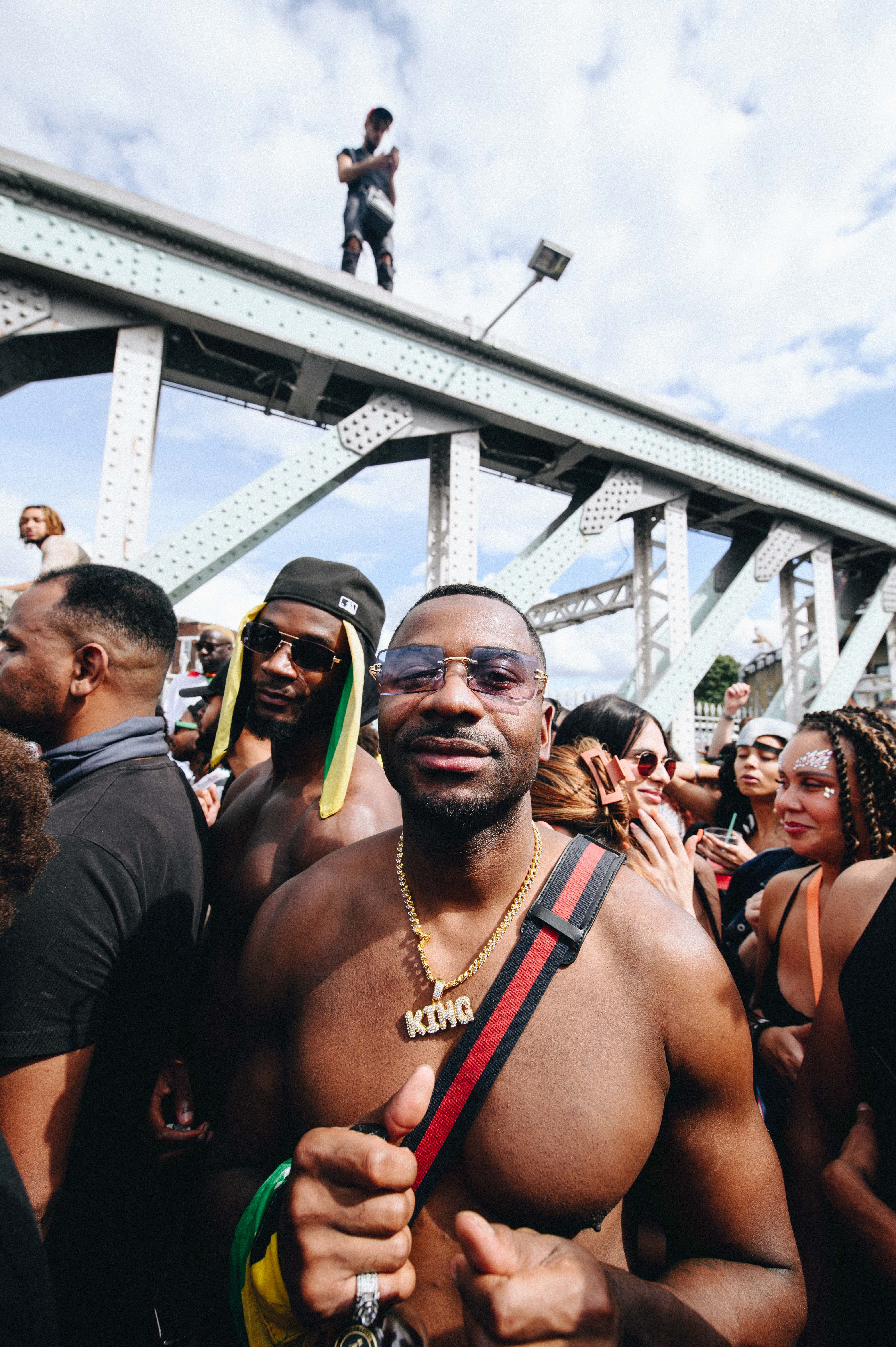 Notting Hill Carnival 2023, London: A topless man with a necklace reading KING smiles at the camera