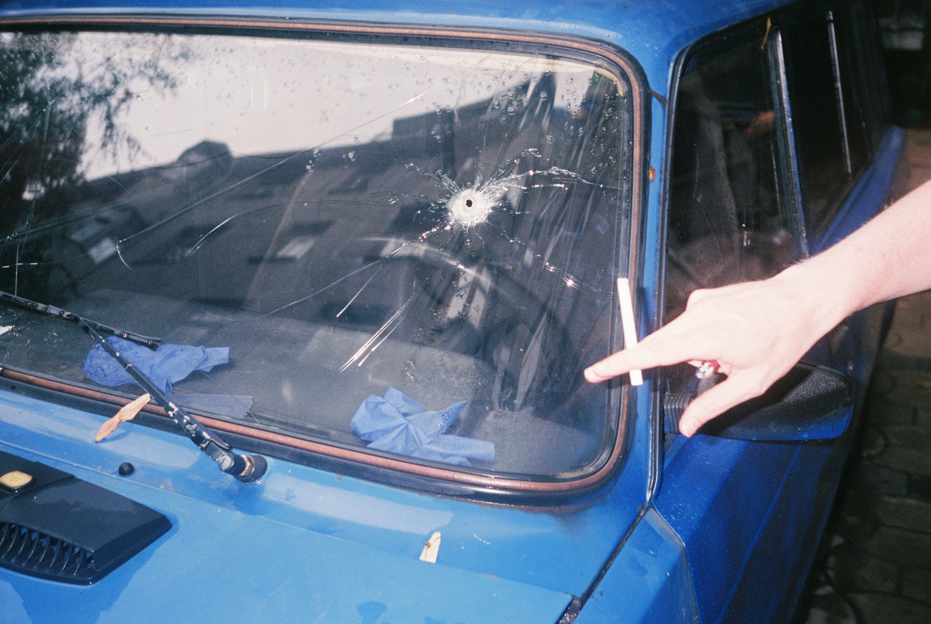 Someone holding a cigarette points at a bullet hole in the car windsheild