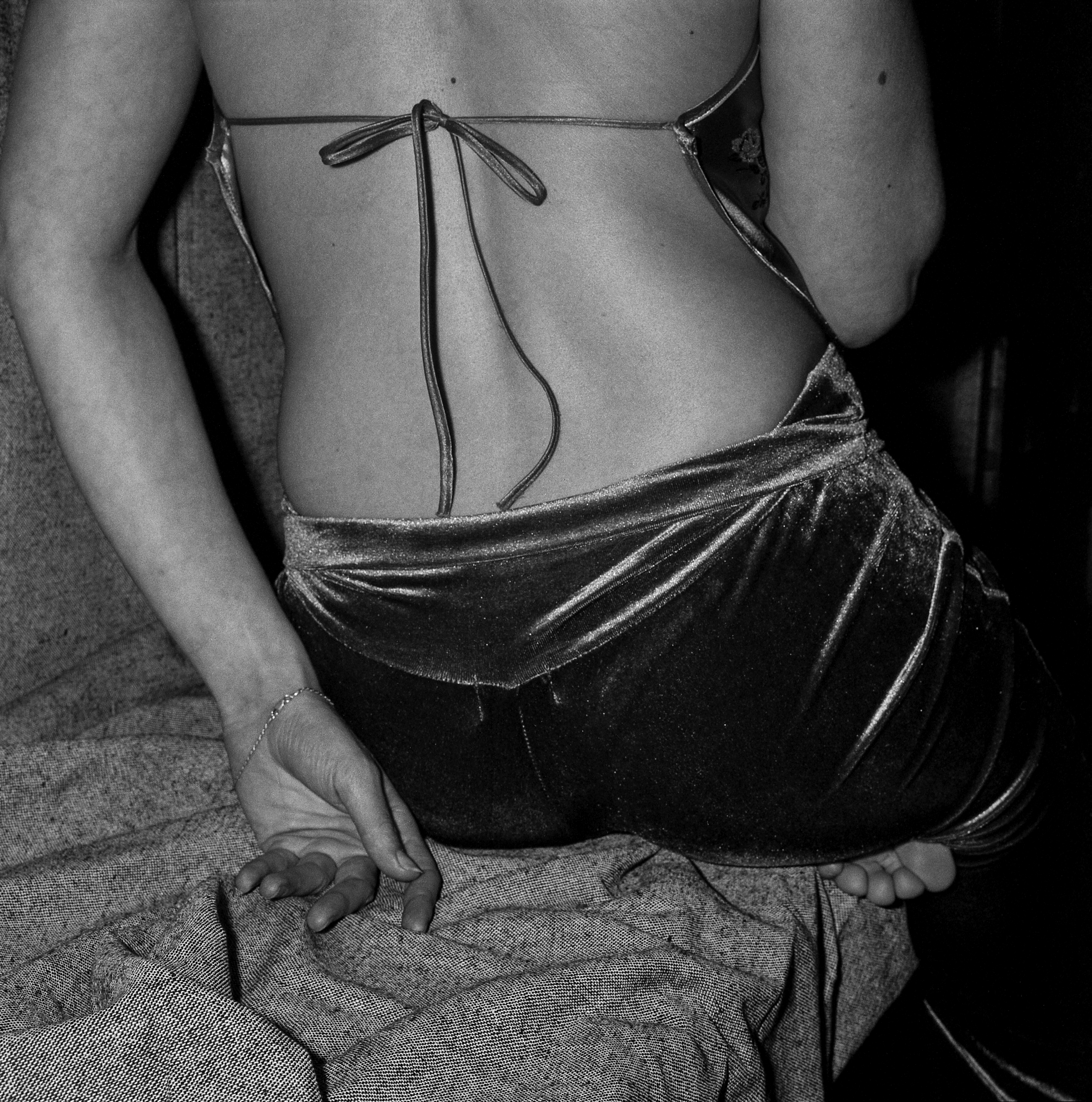 A close up photo of someone wearing a backless velour dress, sitting on a couch, their left arm slouching behind them.